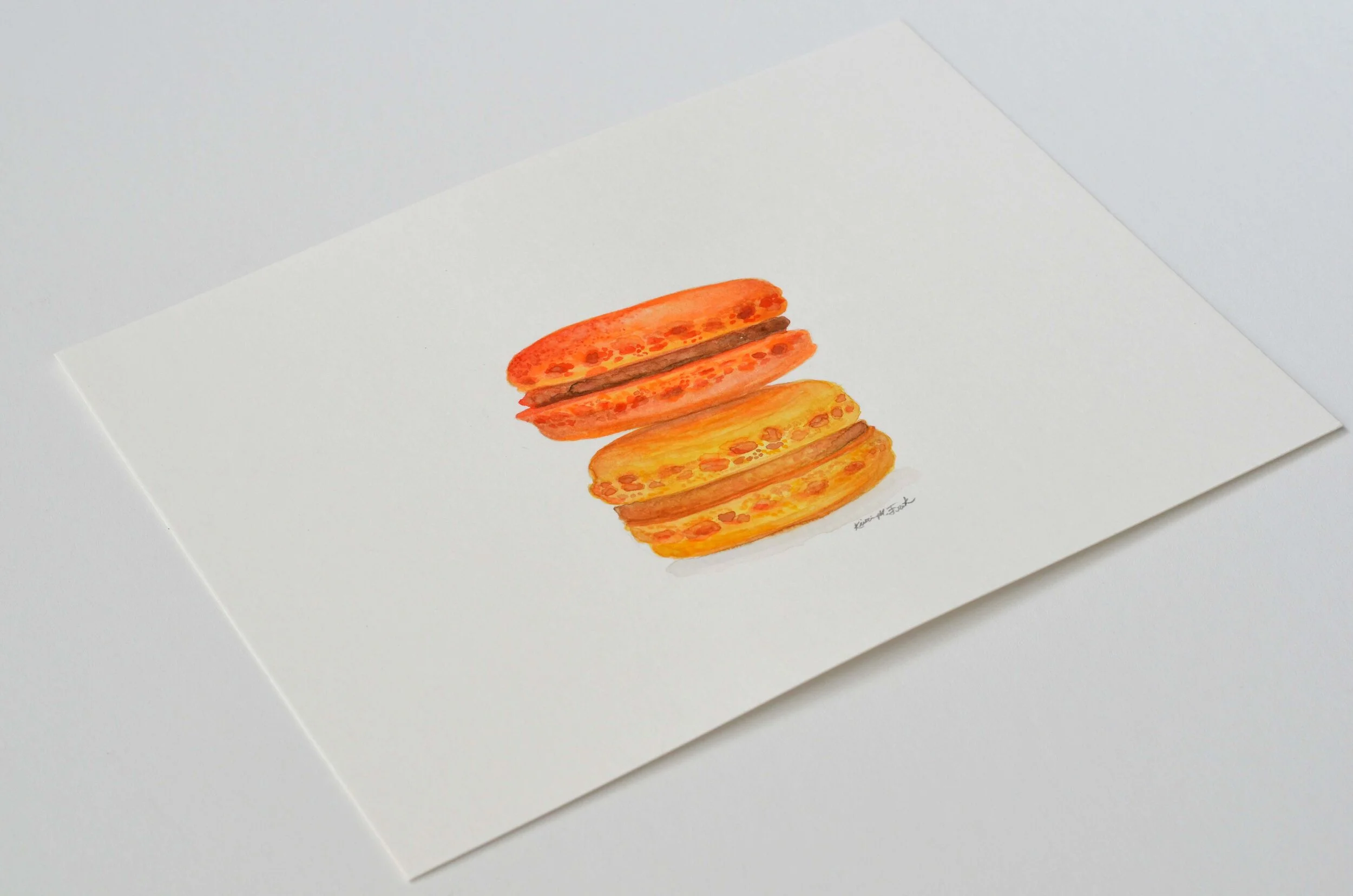 Photo of the watercolor titled "Macaron Monday No.1" by Kristin Frank. The painting features a detailed rendering of two macarons stacked on top of each other. One is orange and one is yellow.