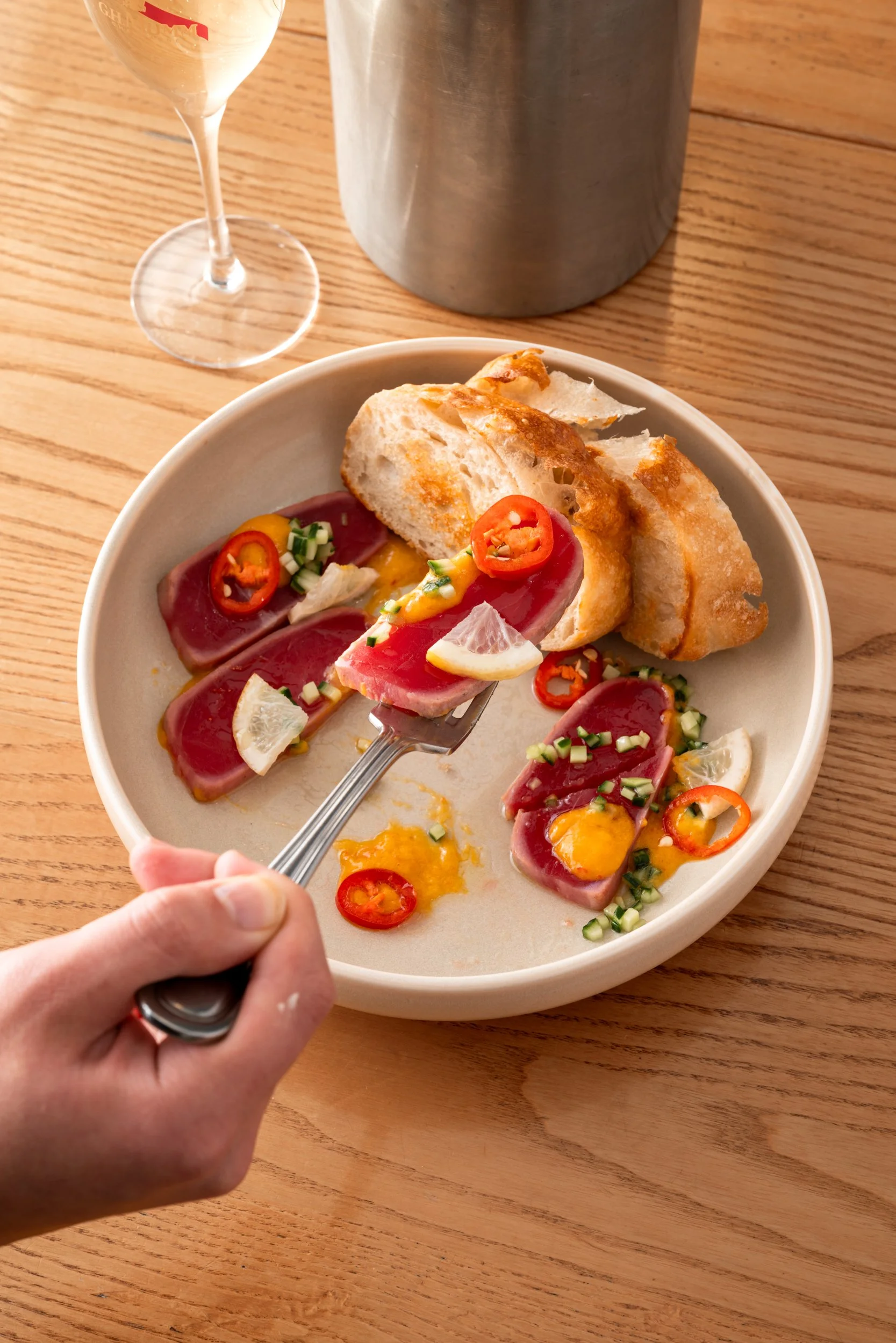 A plate with slices of seared tuna topped with chili peppers, chopped green onions, and lemon wedges, accompanied by pieces of toasted bread, on a wooden table. A glass of white wine and a metal container are in the background.