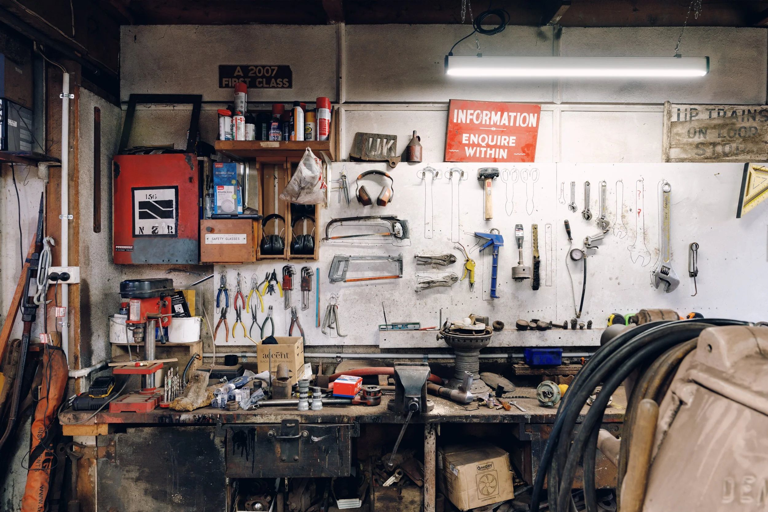 A cluttered woodworking or mechanic workshop with tools hanging on a white wall, including wrenches, pliers, saws, and a hammer. There are shelves with cans of spray and safety glasses. The workbench below holds various small tools and parts. Signs o