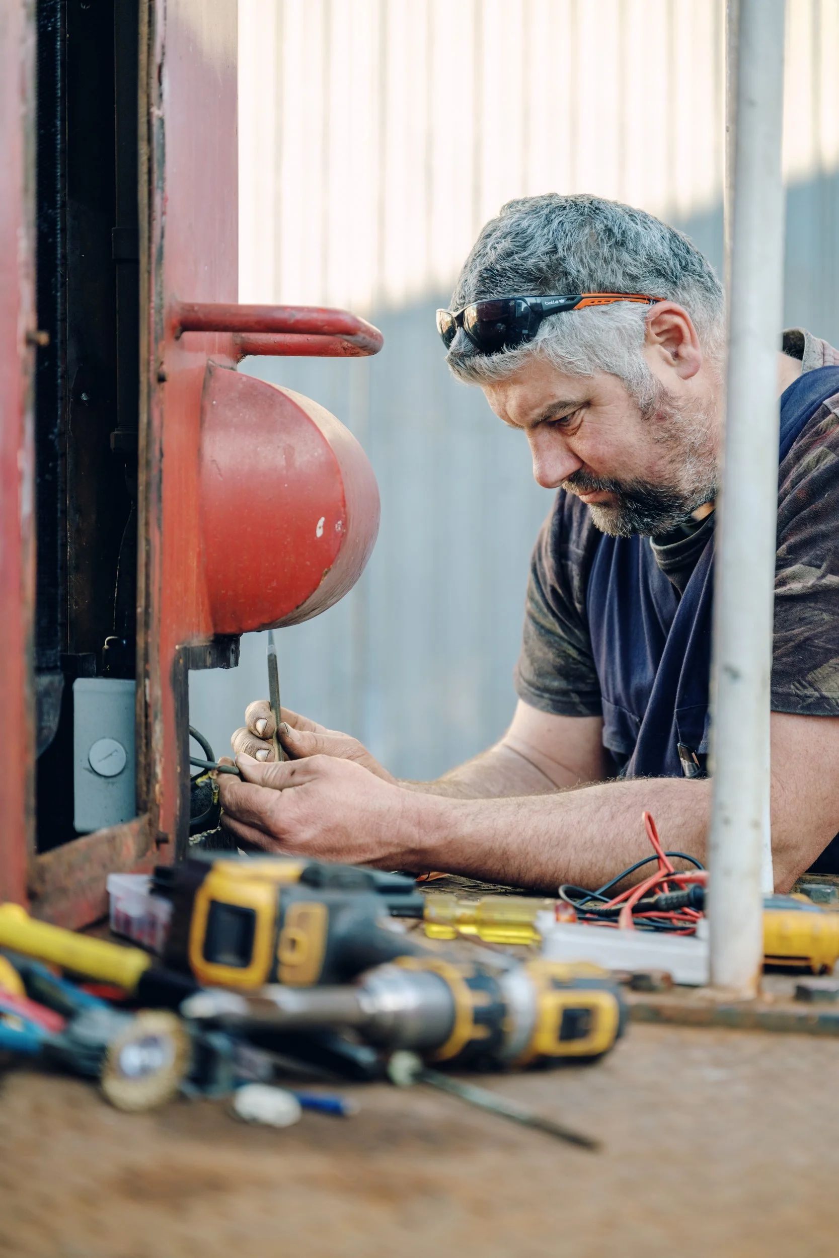 A man with sunglasses and gray hair is fixing or working on an electrical or mechanical device at a workbench outdoors, with tools and wires scattered around.