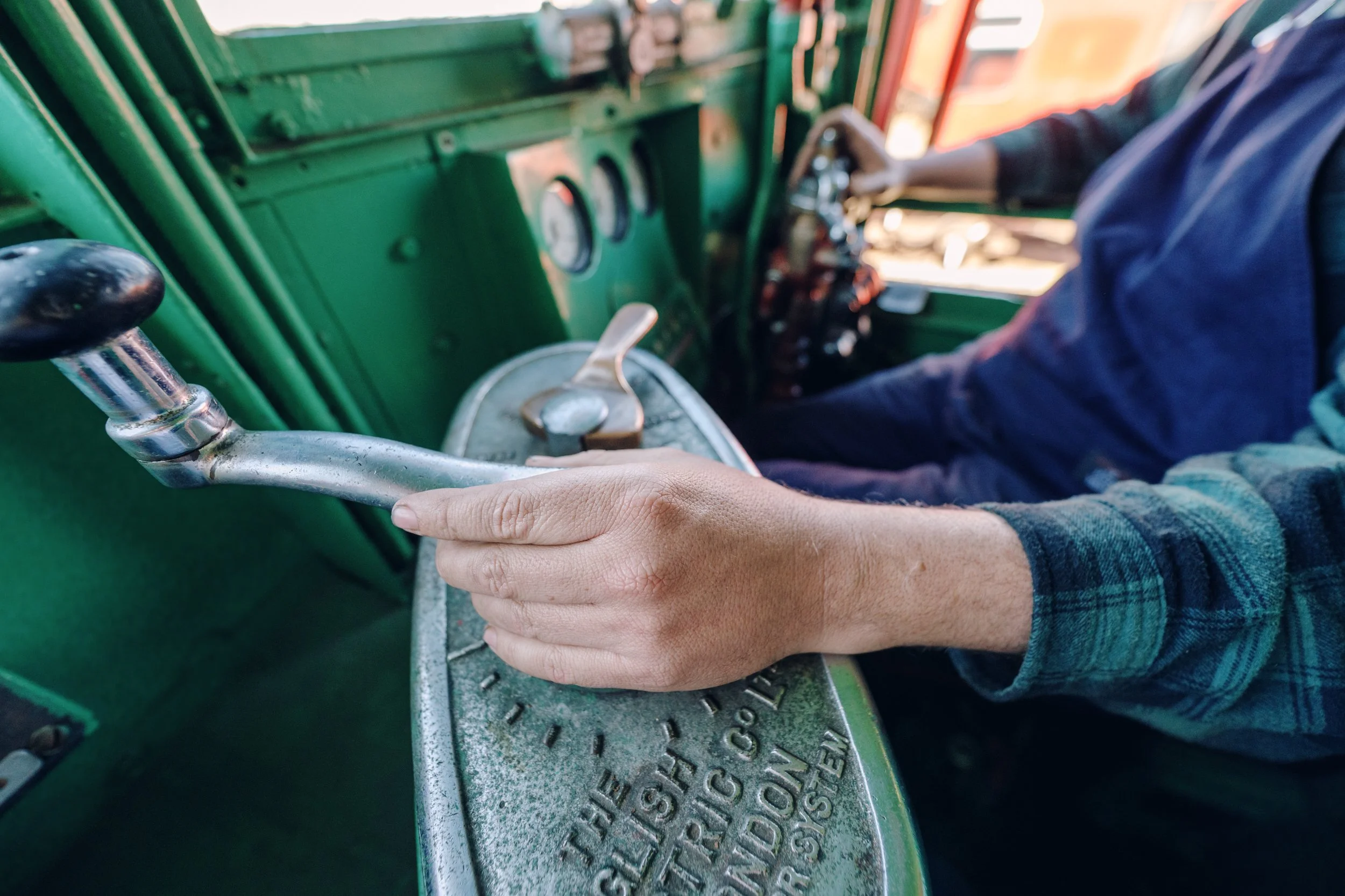 Close-up of a person operating a vintage green truck, with their hand on the gear shift and dashboard gauges visible.