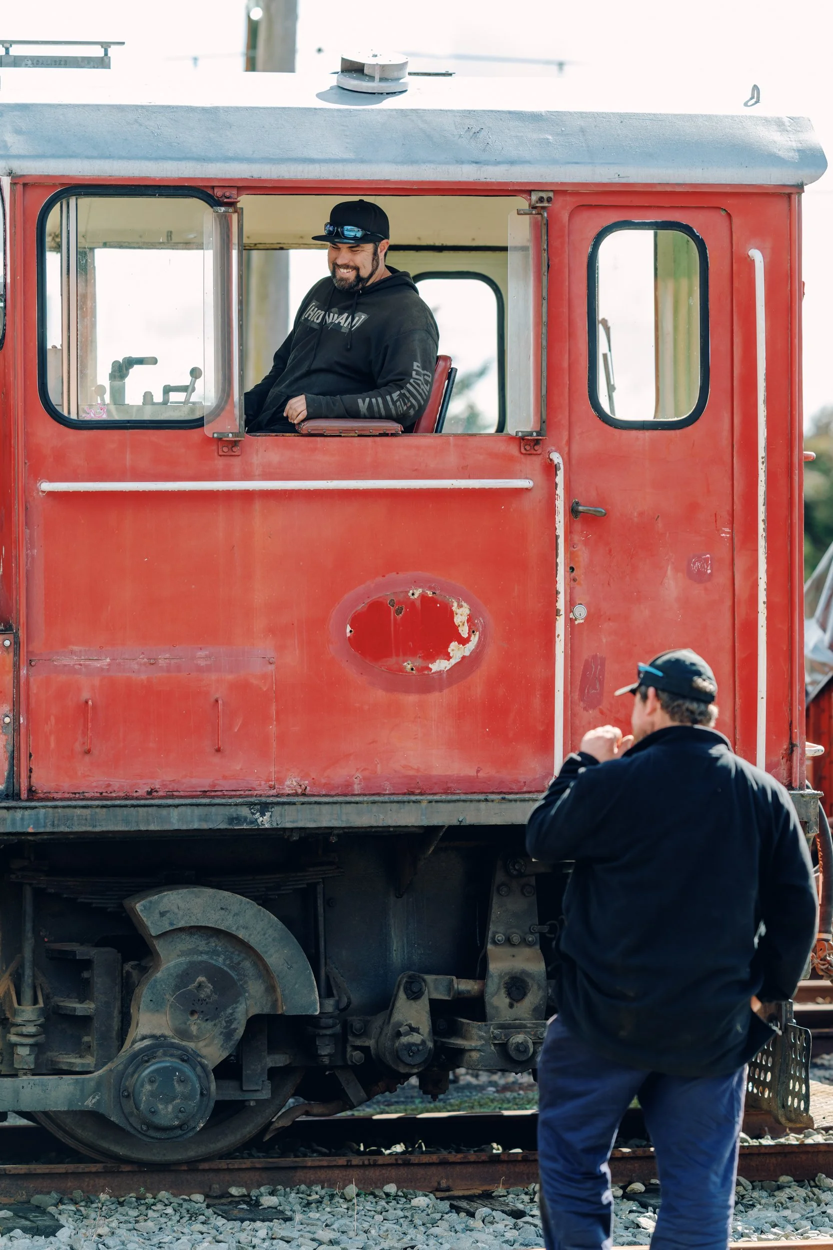 Two men and a red train car. One man is sitting in the train cab, smiling, wearing a black hoodie and a cap with sunglasses. The other man is standing outside on the railroad tracks, facing the train, wearing a black jacket and a cap.