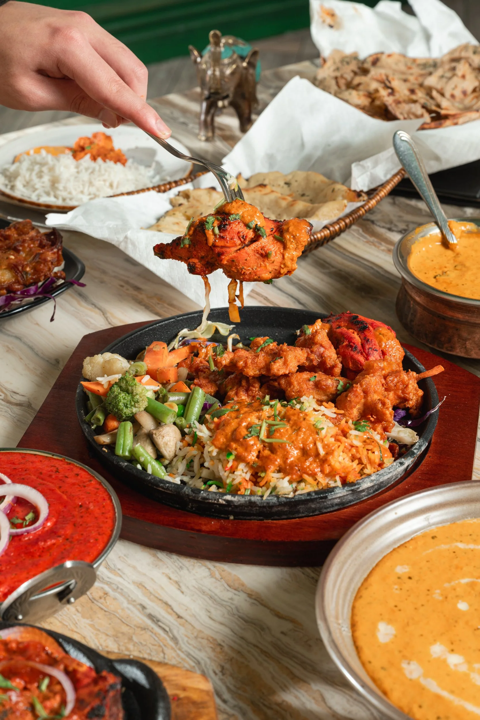 A hand holding a spoonful of chicken tikka masala over a hot plate of Indian food including rice, vegetables, and sauces, surrounded by other dishes on a marble table.