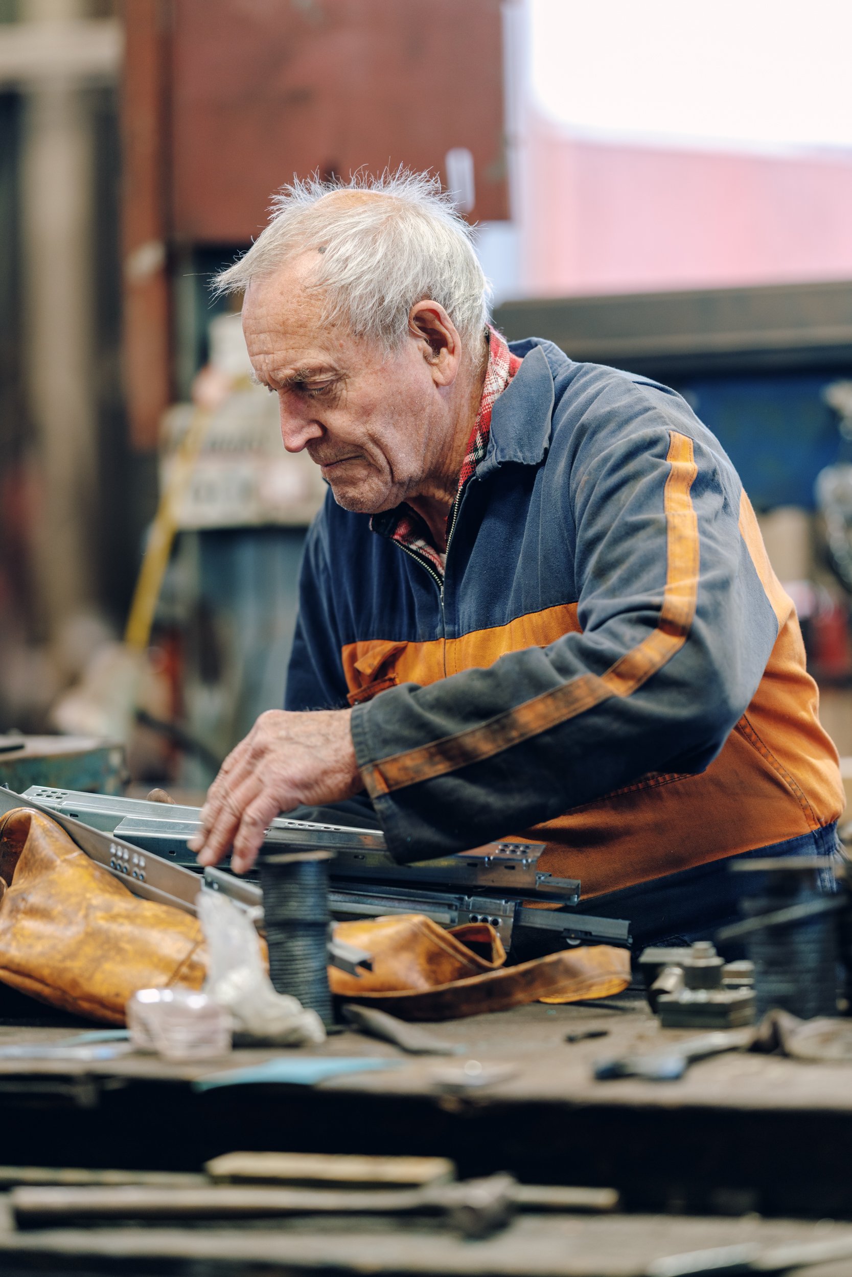 An elderly man with gray hair working on a metal piece in a workshop, surrounded by tools and equipment.