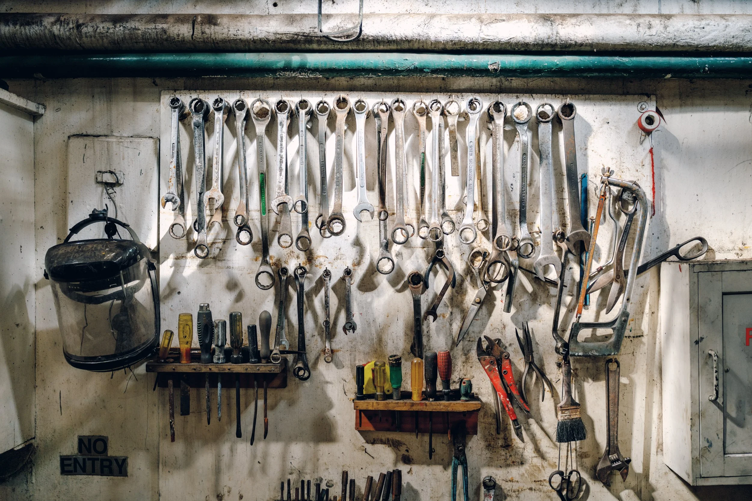Wall with tools hanging, including wrenches, pliers, screwdrivers, and a welding helmet, with a small cabinet to the right and a 'No Entry' sign on the lower left.