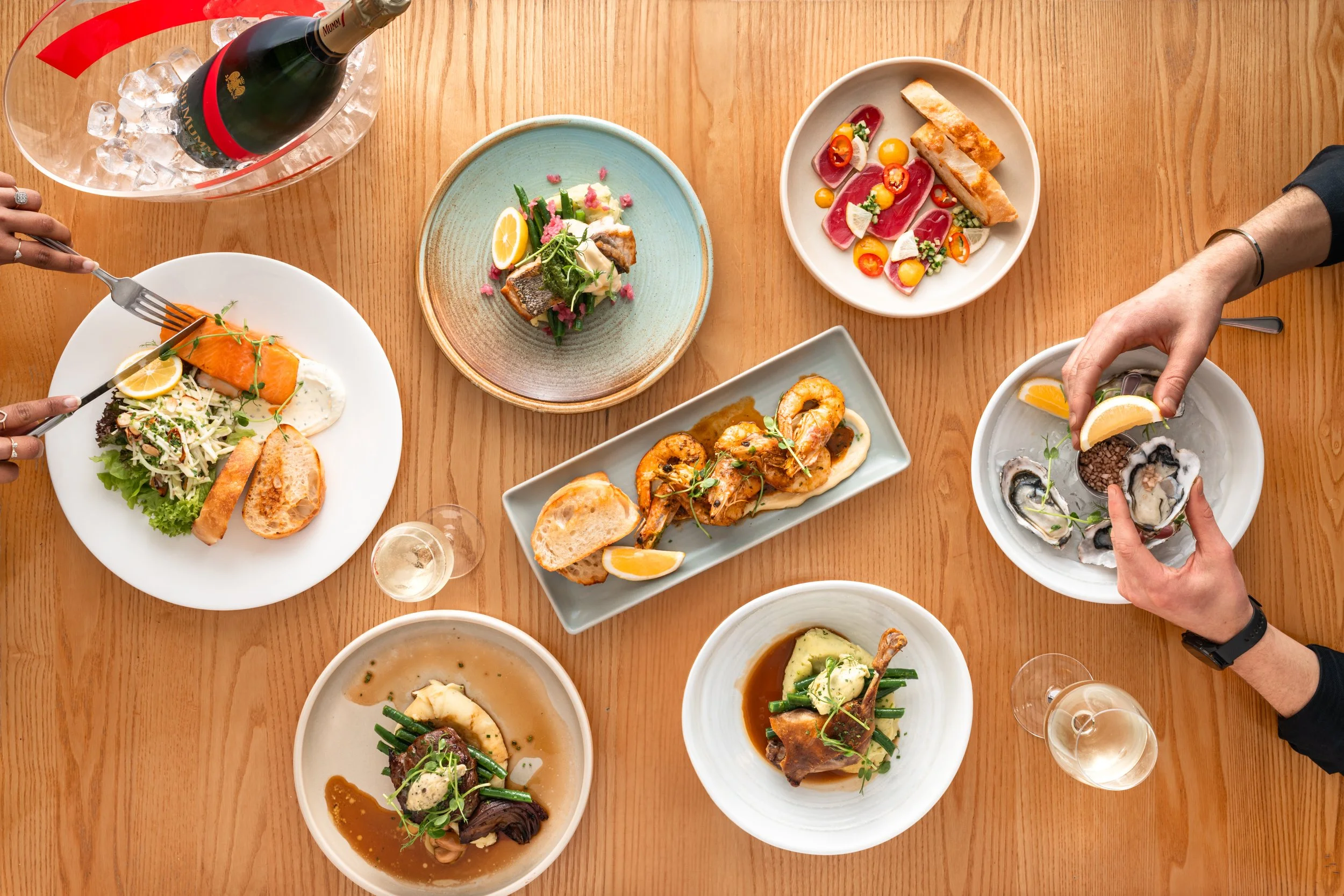 A wooden table filled with various plates of gourmet food, including seafood, meat, vegetables, and a bottle of champagne with glasses, seen from above.