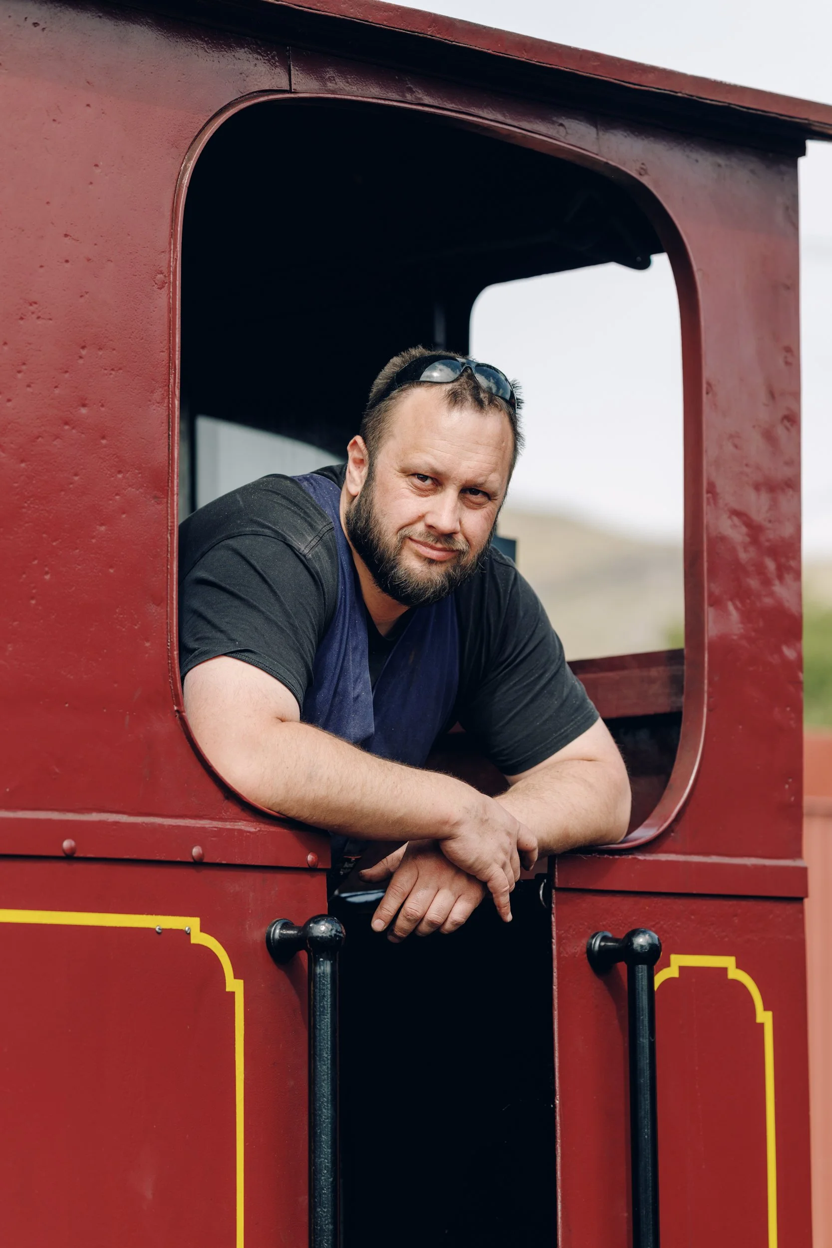 A man with a beard and sunglasses on his head leaning out of a red train's window, outdoors with a blurry background of hills.