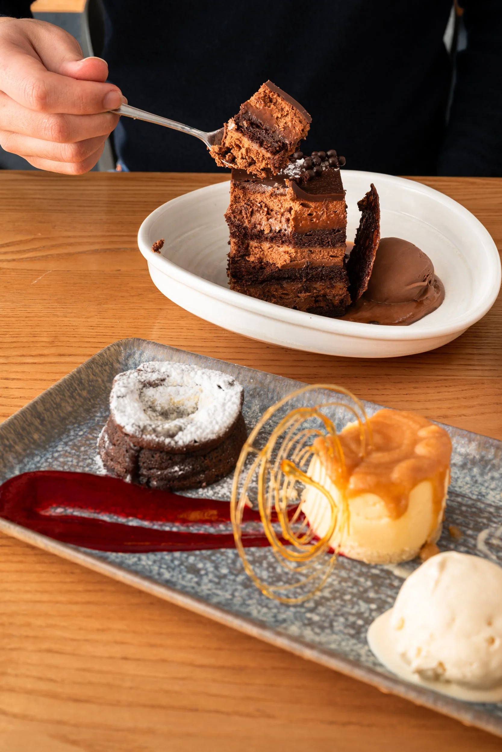 A person holding a fork with a bite-sized piece of layered chocolate cake, with a white oval plate of a larger slice of the same cake on a wooden table. In the foreground, a rectangular ceramic plate holds a chocolate lava cake dusted with powdered s