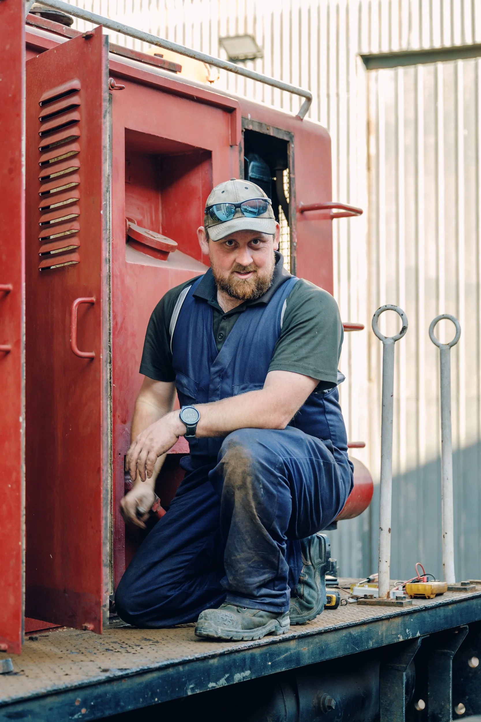 A man with a beard, wearing a cap, sunglasses, and workwear, kneeling on a platform next to a red fire truck, working on equipment.