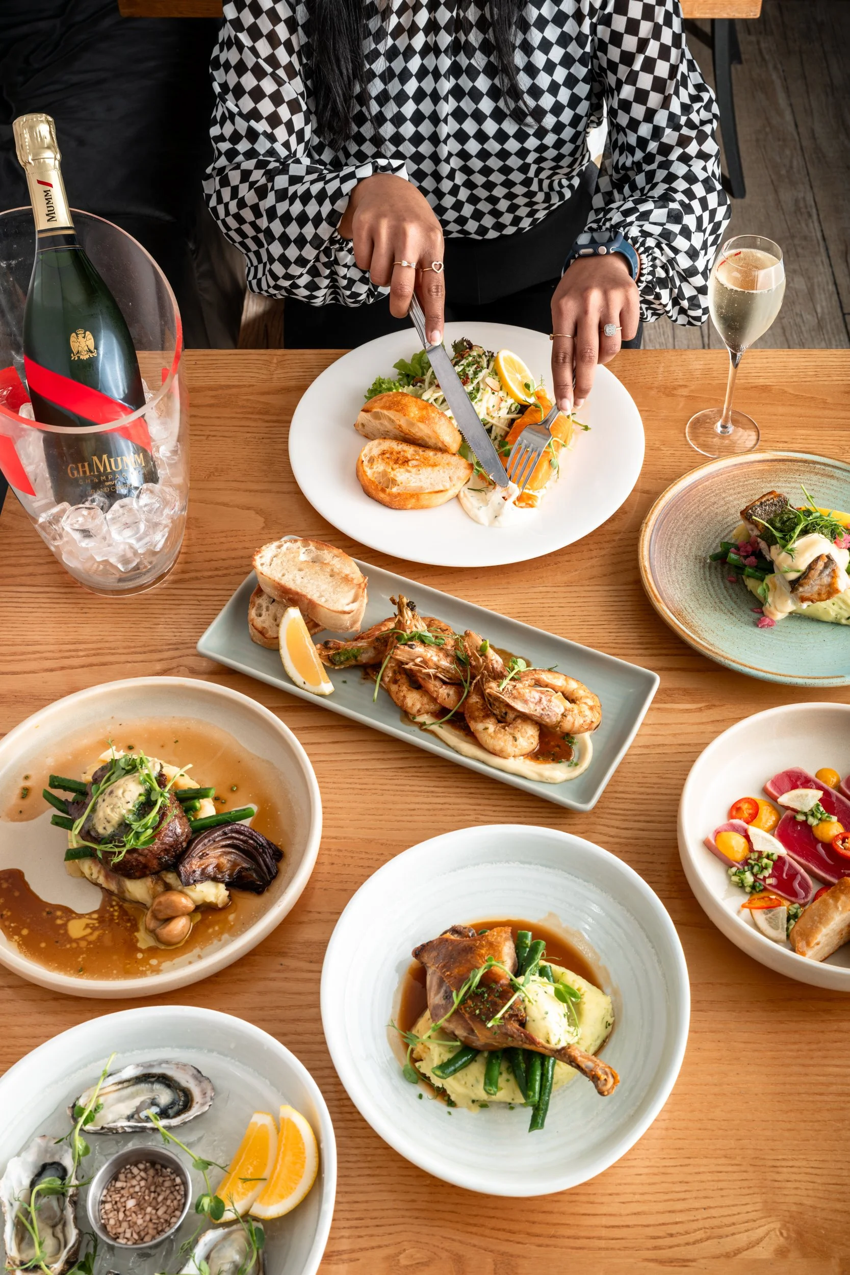 A person in a checkered shirt cuts into a salad on a white plate at a table with multiple gourmet dishes, a bottle of champagne in a bucket, and glasses of champagne.