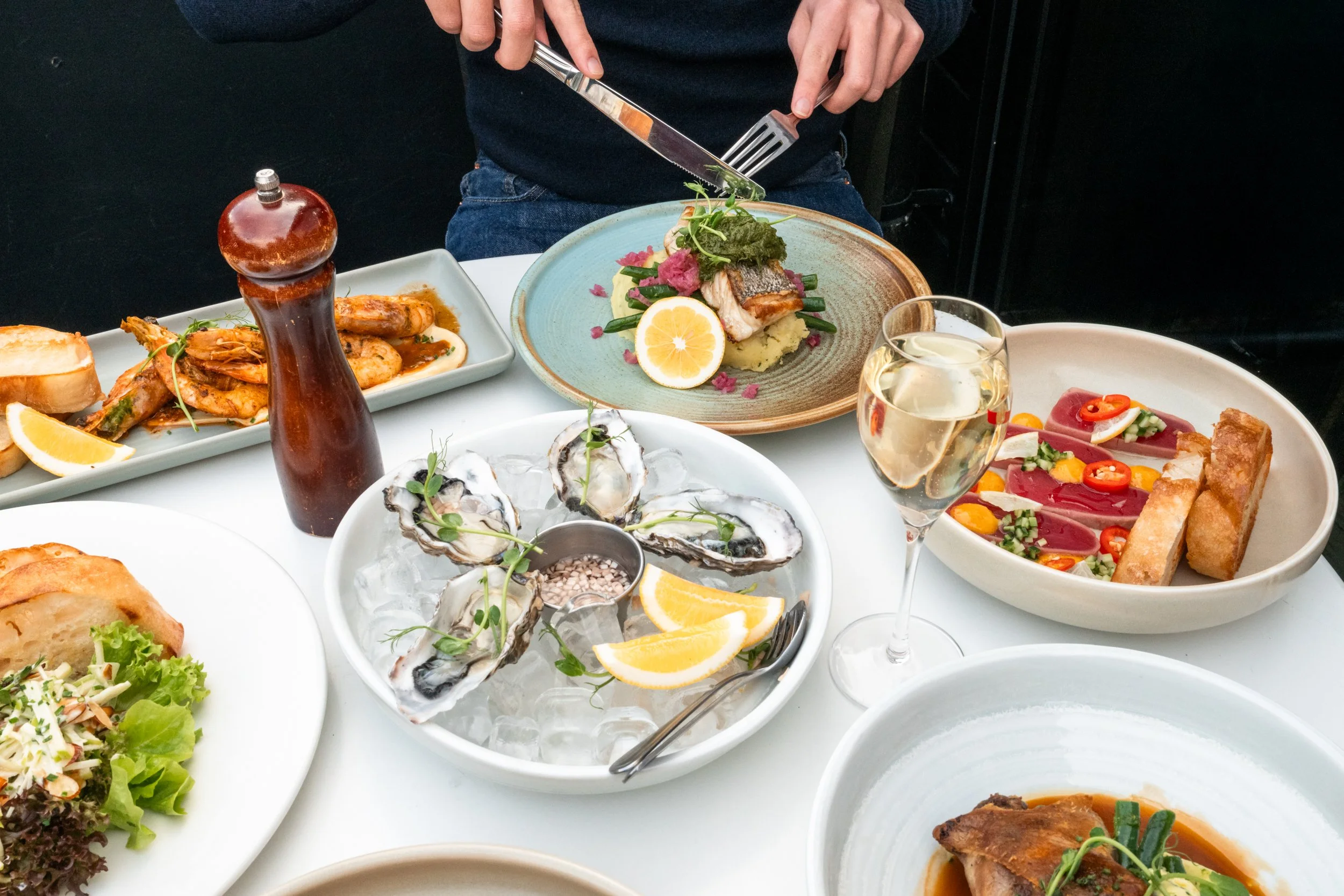 Assorted dishes including oysters on ice, a seafood platter, a plate of sliced fish with lemon, a glass of white wine, a salad, and a plate of shrimp and chicken, along with a person preparing food.