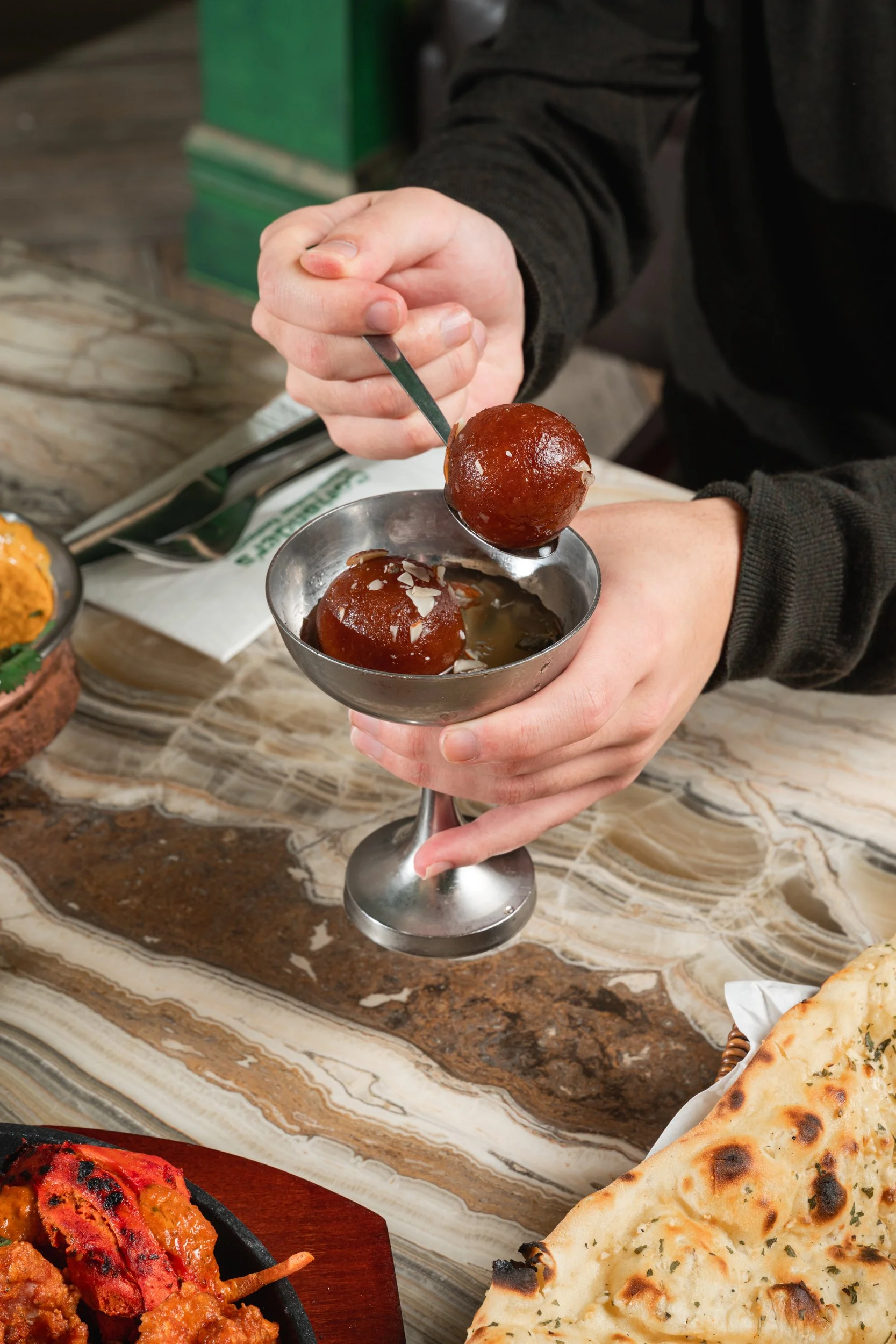 Person holding a metal bowl with two gulab jamuns, one on a spoon above the bowl, drizzling syrup over the dessert.