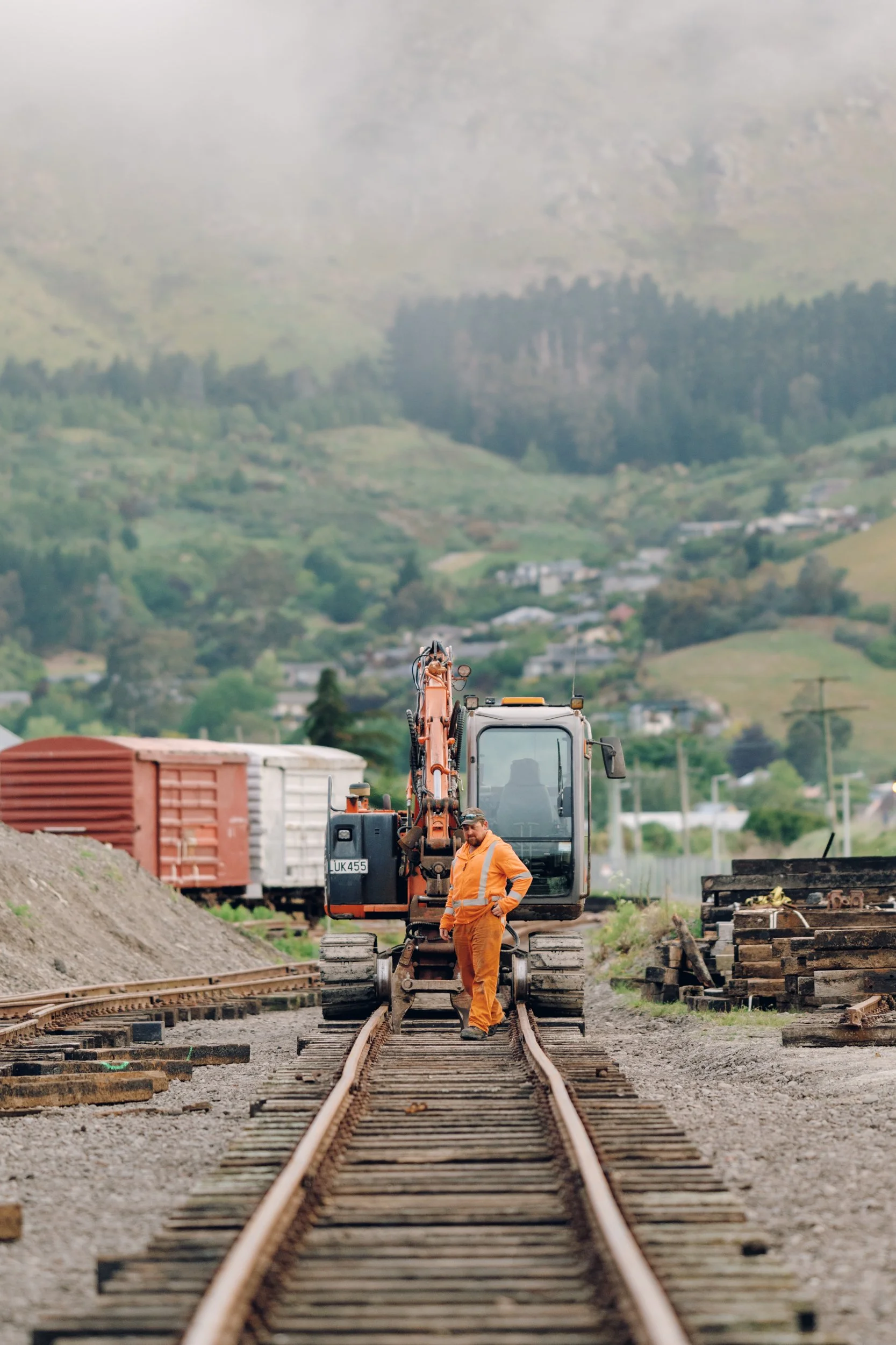 A construction worker in orange safety gear standing on railway tracks with construction equipment and train cars in the background, surrounded by hilly green landscape.