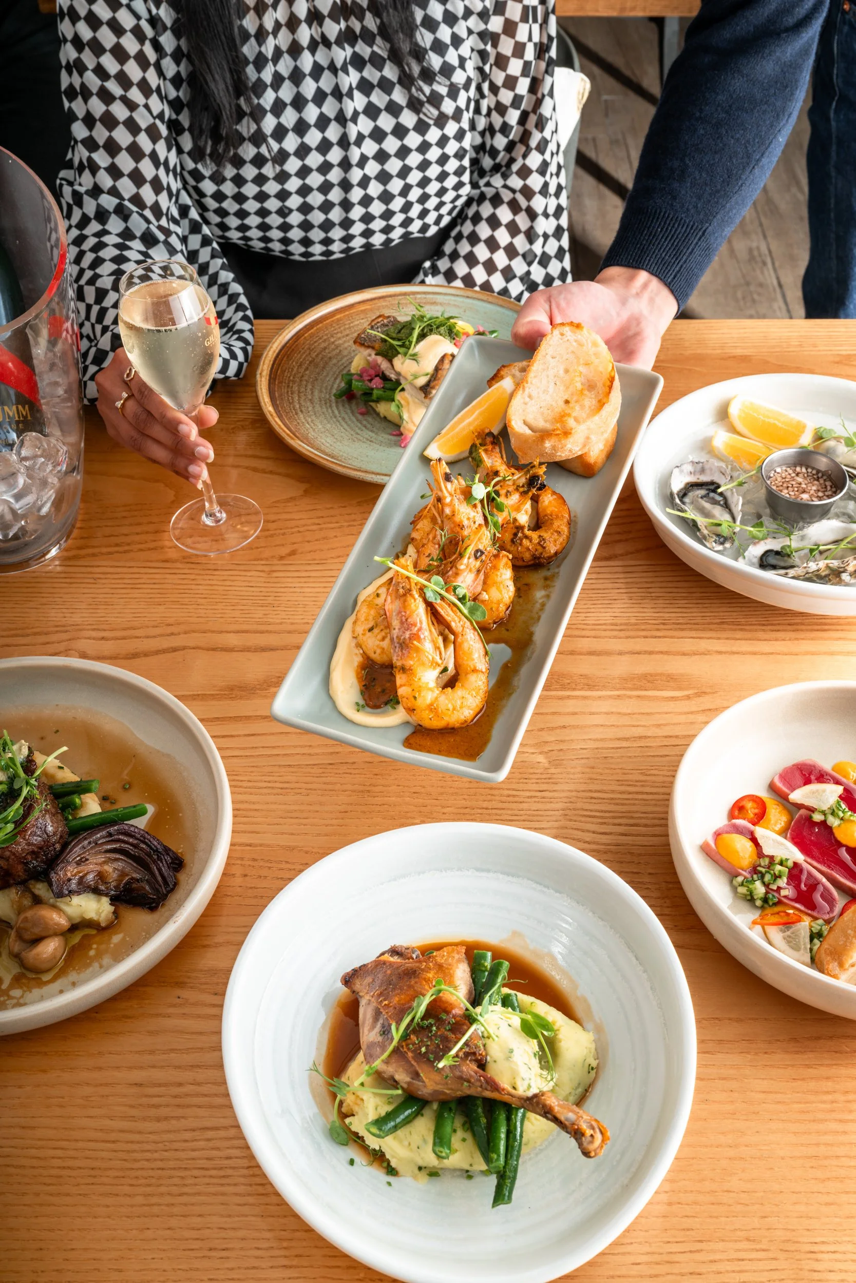 A person holding a plate of shrimp with lemon and bread, surrounded by various dishes including salad, roasted chicken, oysters, and beef with vegetables, on a wooden table, with a glass of white wine and a bucket of ice nearby.