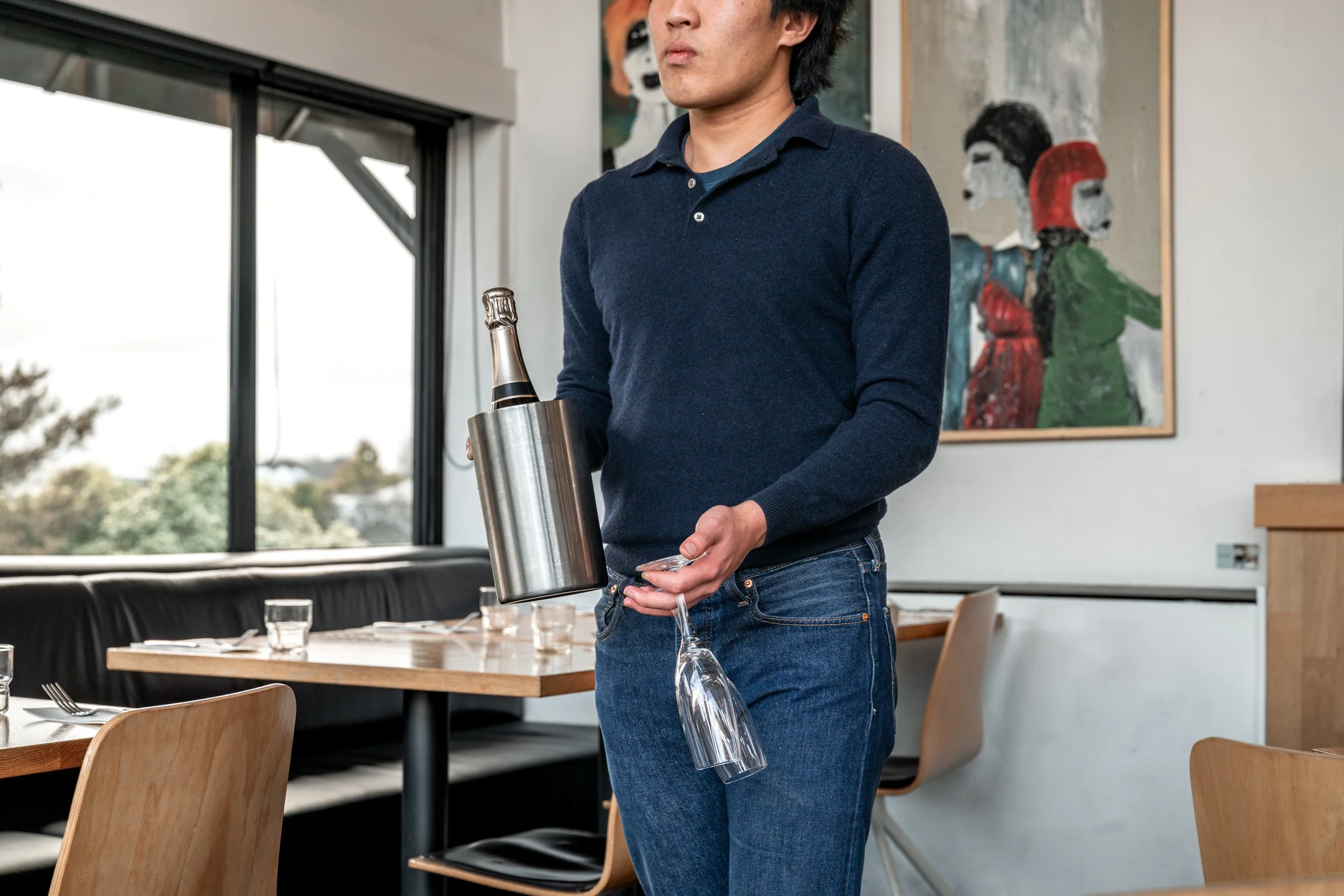 A man in a navy blue sweater and jeans holding a champagne bottle in an ice bucket and a wine glass inside a modern restaurant.