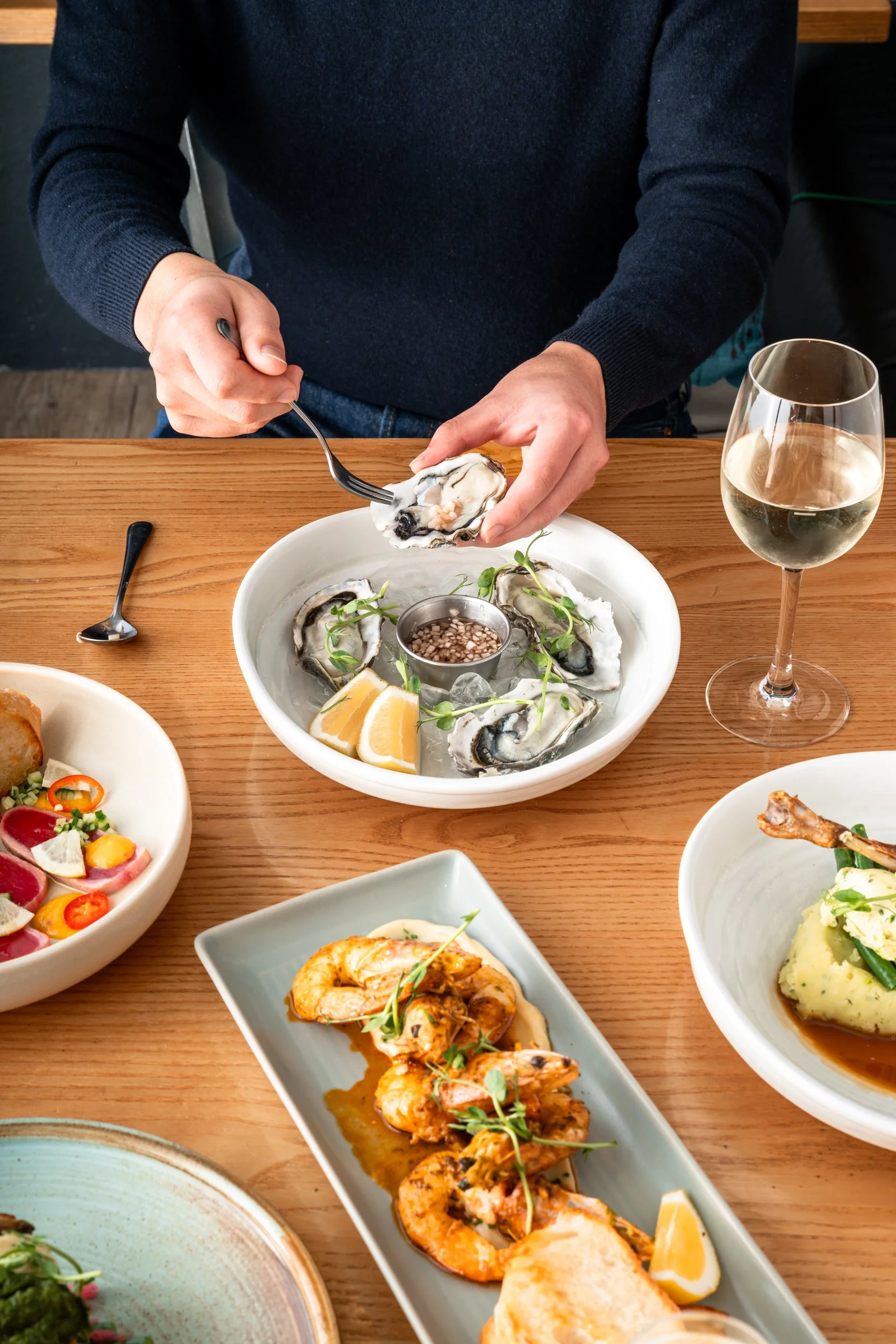 Person wearing a dark sweater eating oysters at a wooden table with various dishes and a glass of white wine.