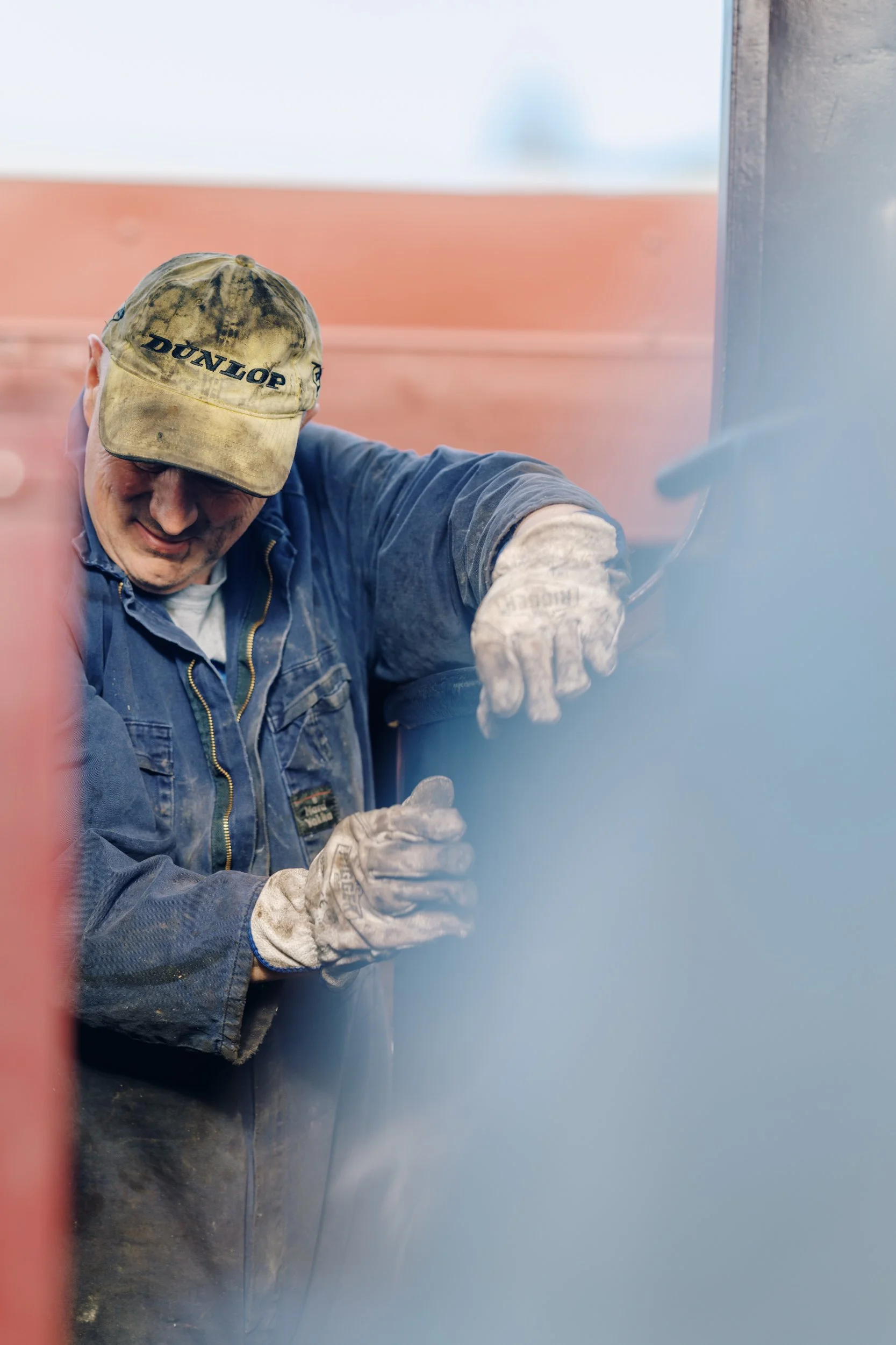 A man wearing a worn baseball cap, a blue work jacket, and gloves is welding or working with metal, focused and smiling.