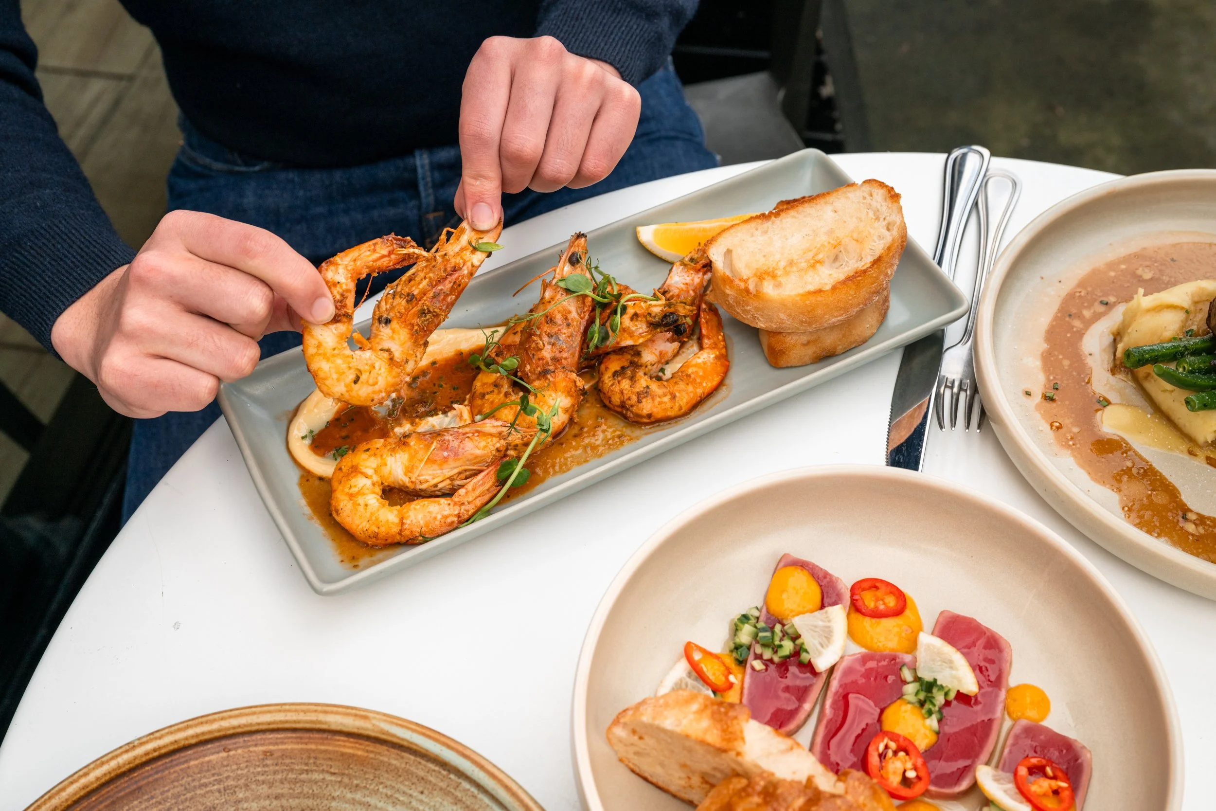 Person reaching to pick up a shrimp from a plate of grilled shrimp in sauce, served with bread on a rectangular plate. Other dishes on the table include a bowl of seared tuna with garnish, and a plate of ravioli with green beans and sauce.