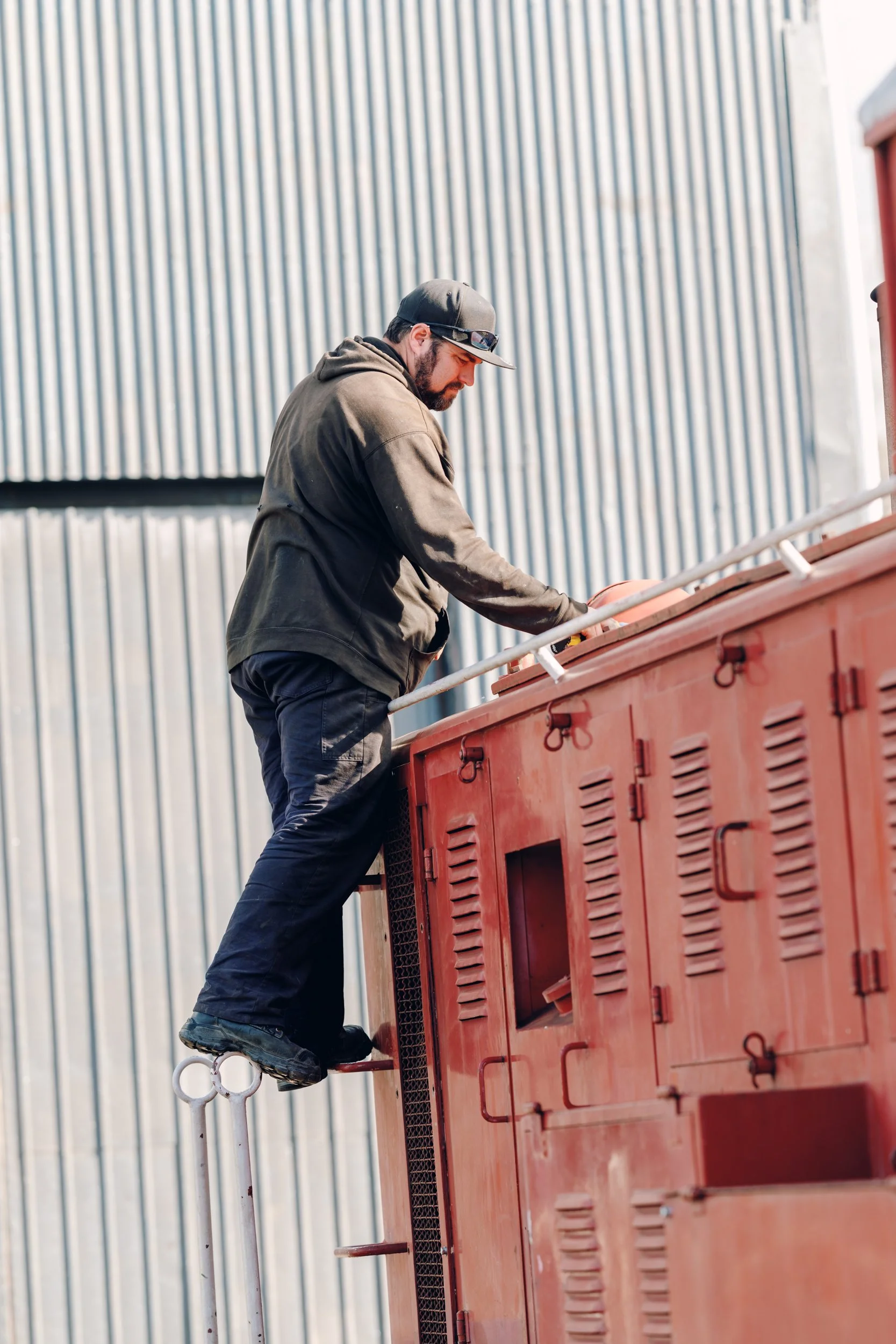 A man wearing a black hoodie, cap, and sunglasses leans over a red industrial machine or container, with a corrugated metal wall in the background.