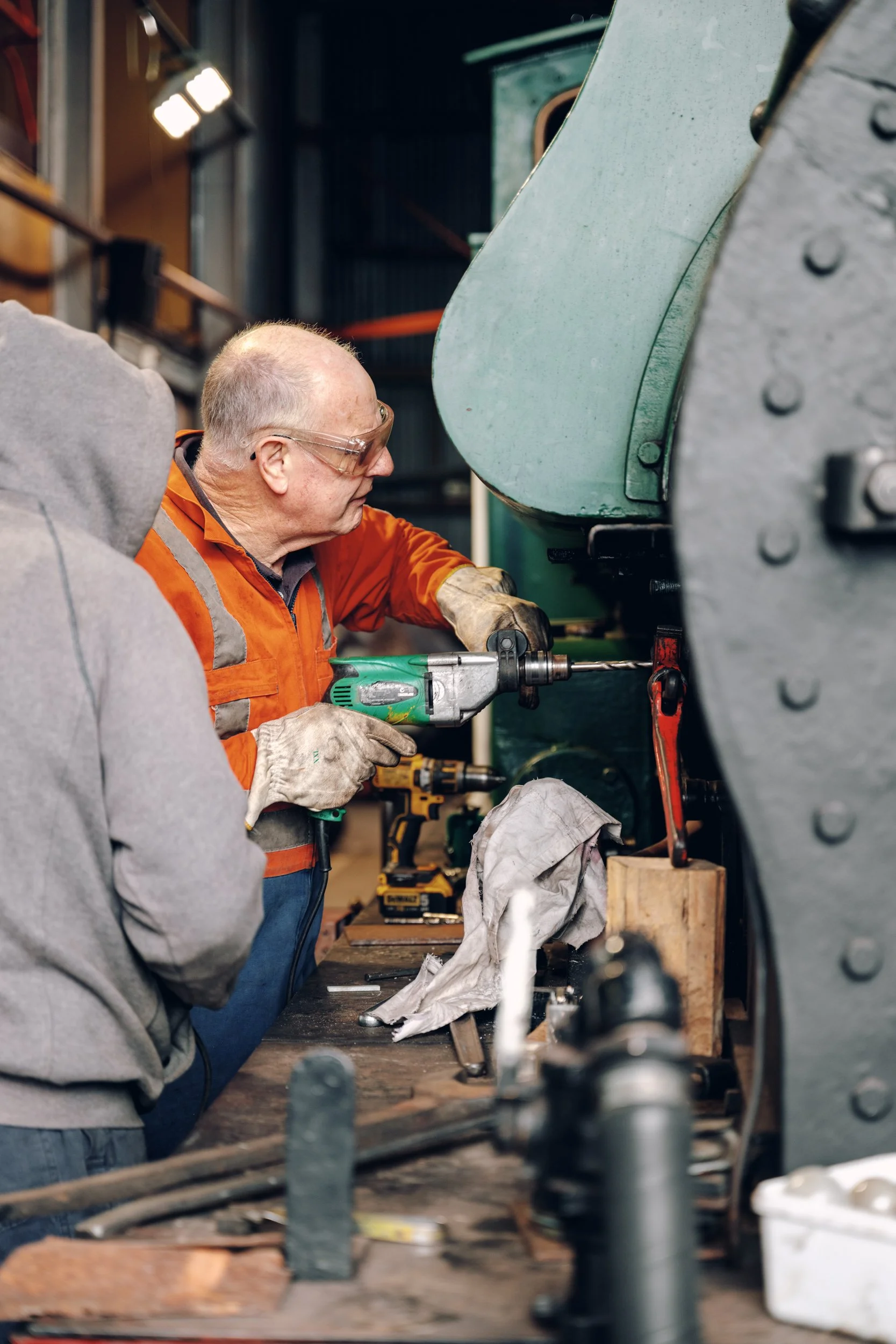 Two men working on a large green machine in a workshop. One man, wearing orange safety gear, glasses, and gloves, is using a power drill, while the other man observes.