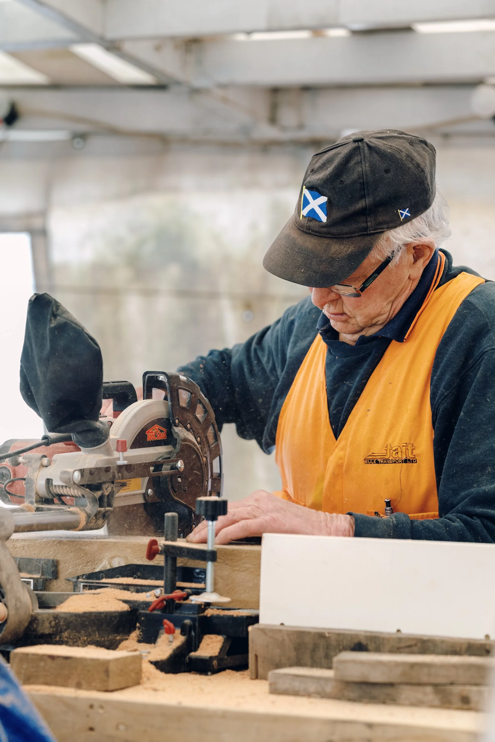 Older man with gray hair wearing glasses, a black cap with the Scottish flag, and an orange vest working with a circular saw in a workshop.
