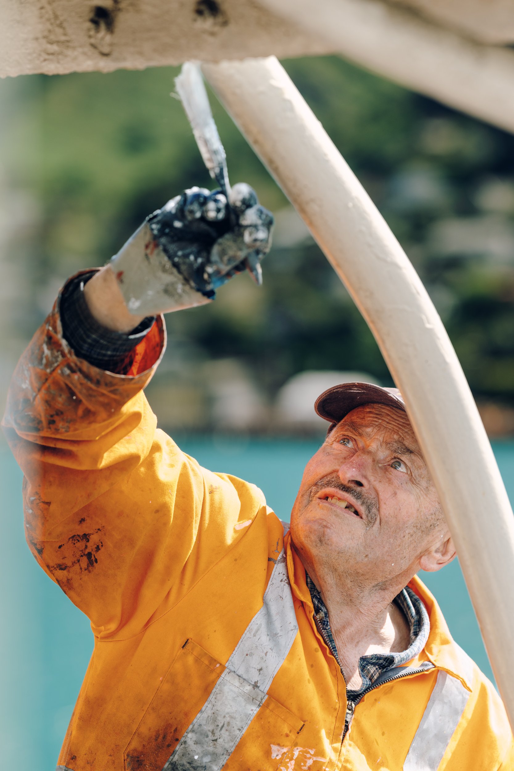 A construction worker wearing a yellow safety jacket and cap painting a large white pipe with a paintbrush.