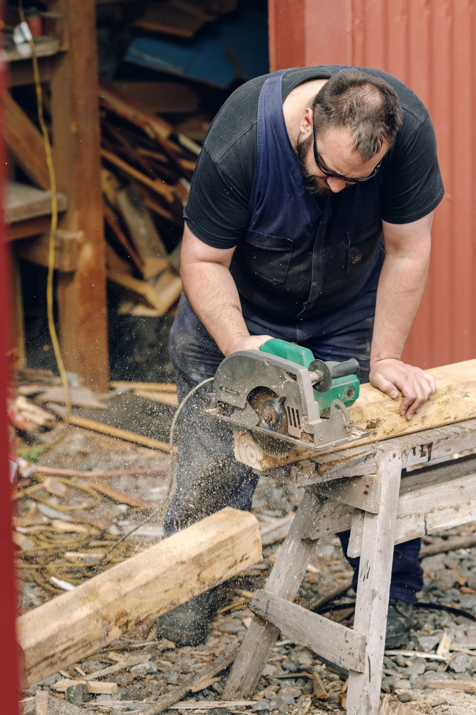 A man wearing safety glasses and a black T-shirt cuts a piece of wood with a green circular saw in a woodworking shop.