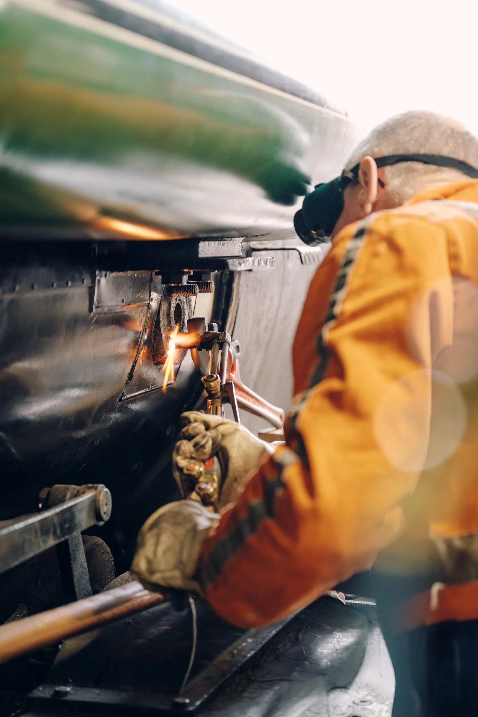 A person welding metal on a ship, wearing safety goggles and a yellow protective jacket.