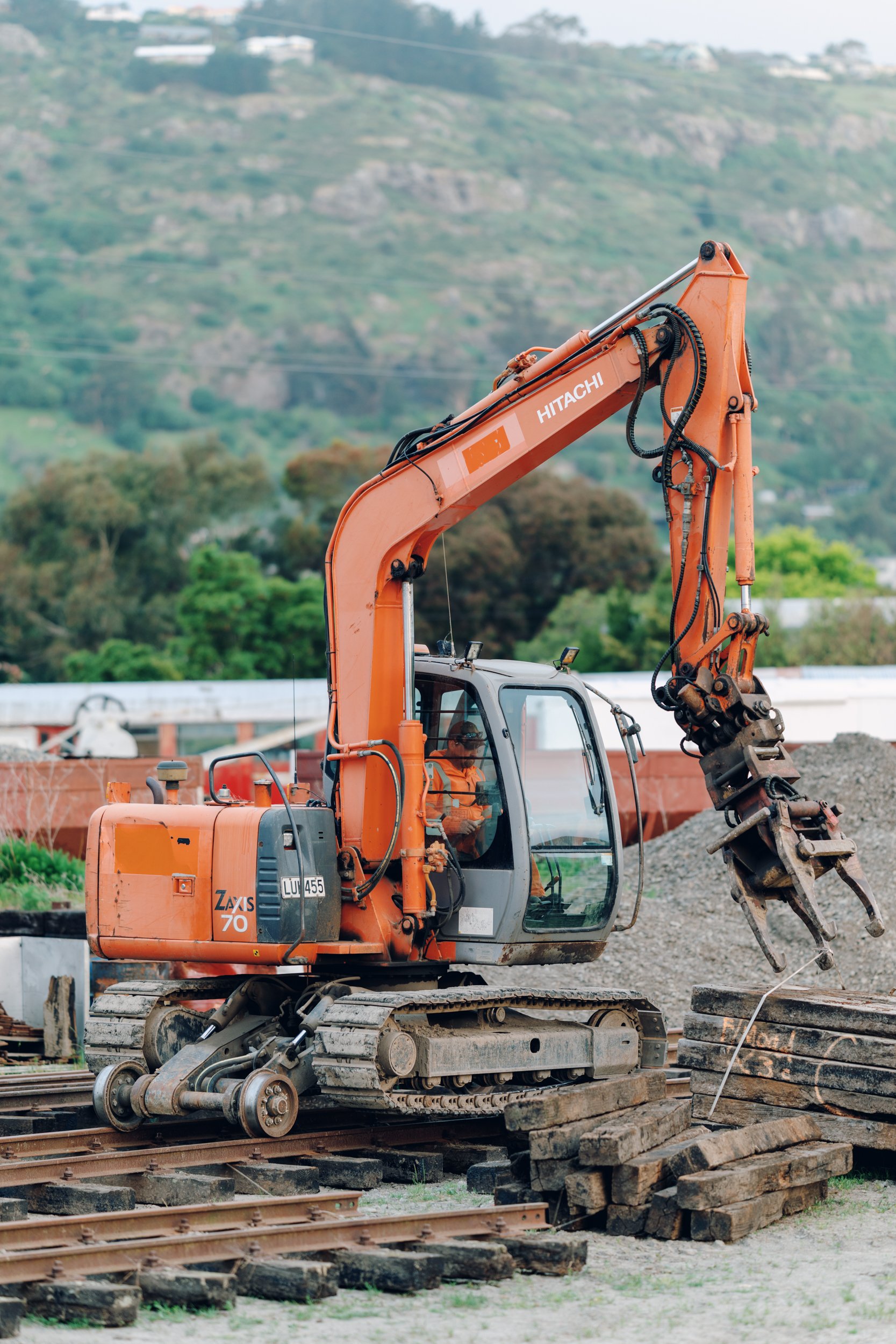 Orange Hitachi excavator working on a construction site with railway tracks, surrounded by green hills.
