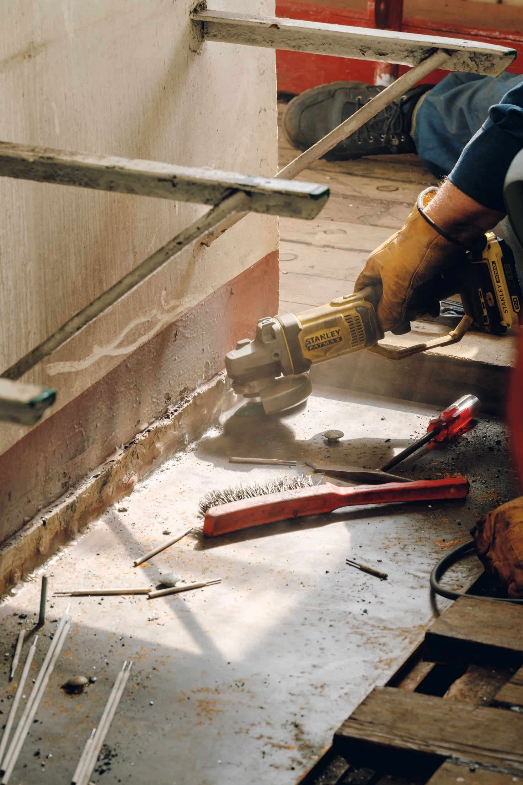A person wearing gloves uses a power tool to grind or cut a concrete floor. Construction tools like a red brush and screwdriver are on the floor, with scaffolding and a wooden pallet nearby.