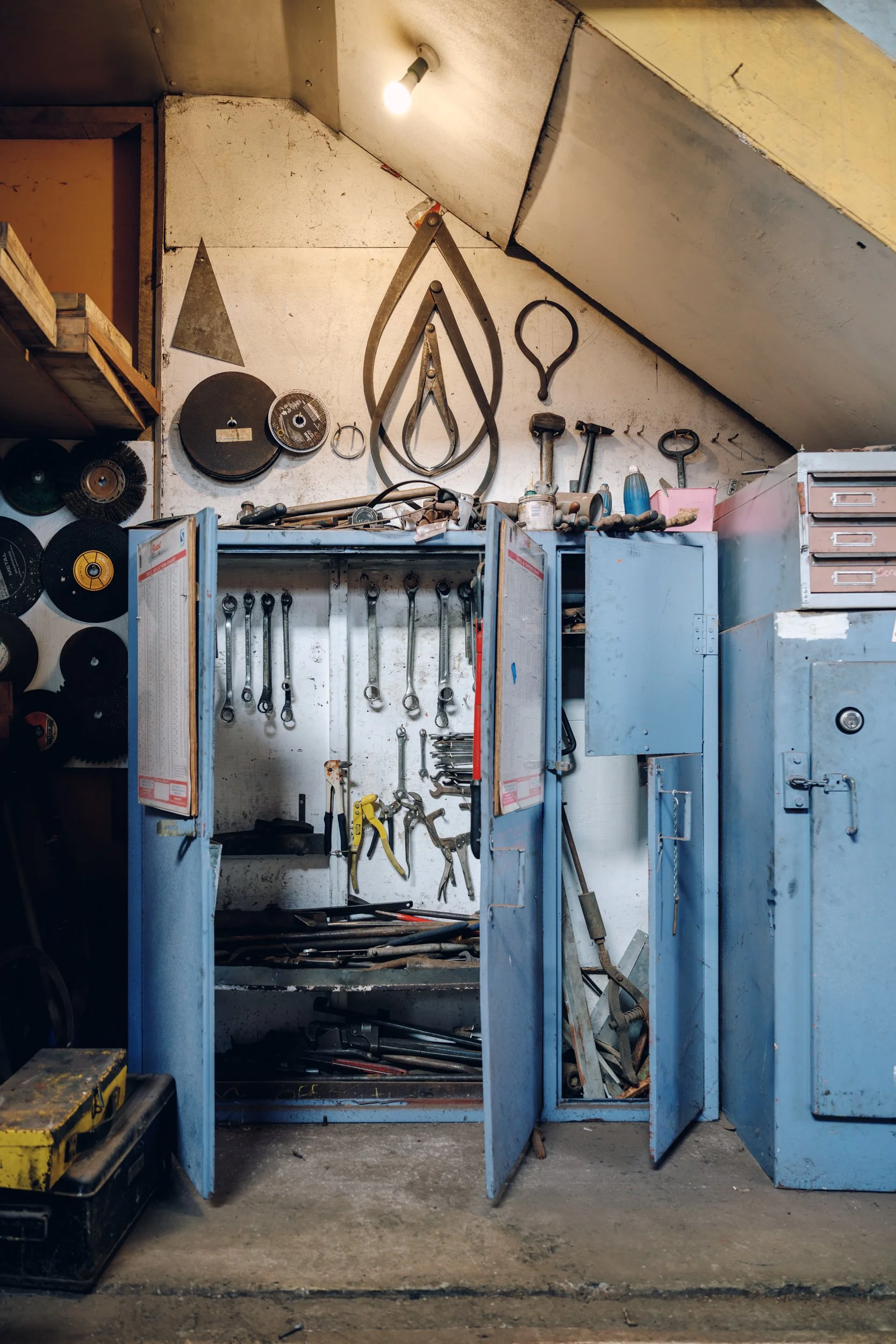 A cluttered workshop with blue tool cabinets, hanging tools, and power tools on shelves, under a sloped ceiling.