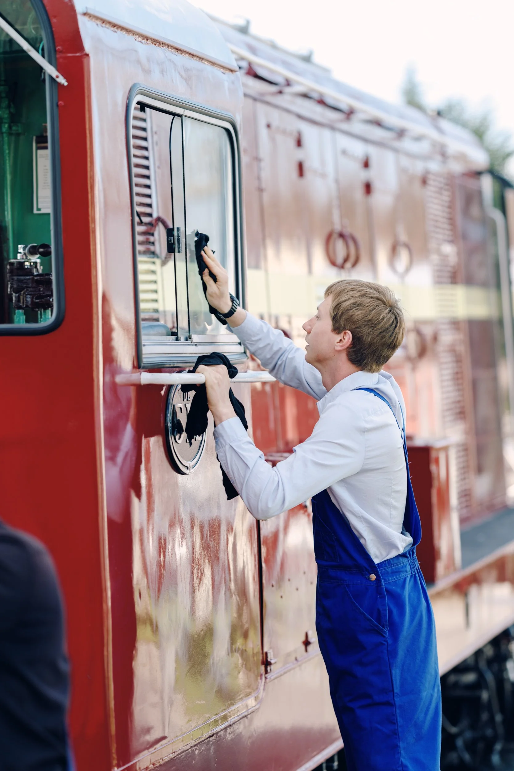 A young man in a white shirt and blue overalls is cleaning the window of a red train with a cloth.