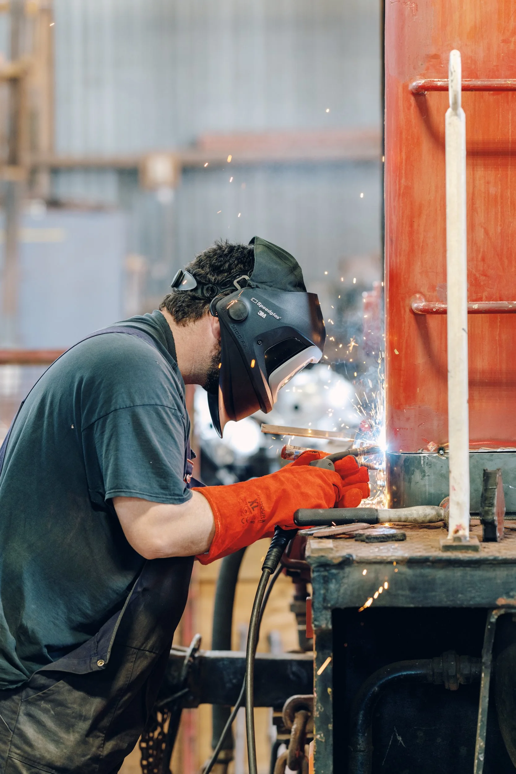 Man welding metal in a workshop wearing safety goggles, a welding helmet, and orange gloves.