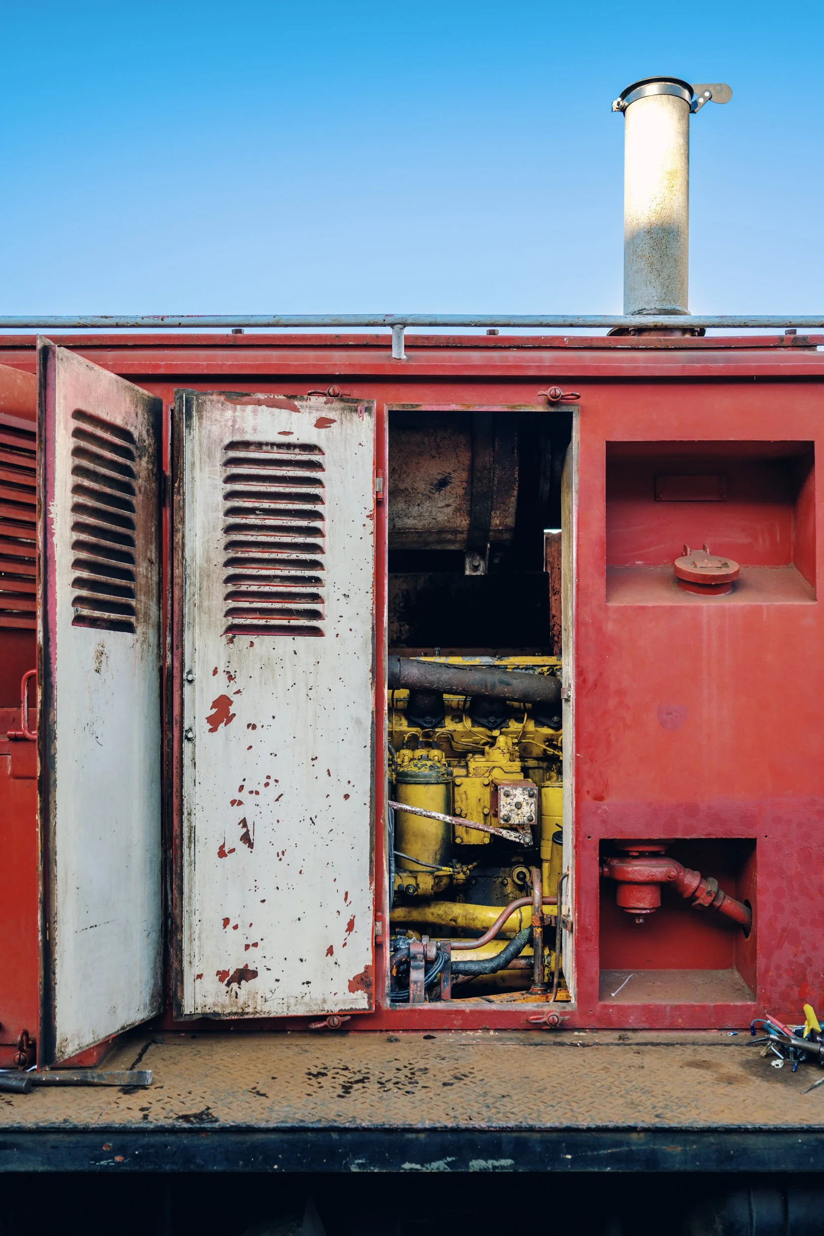 Close-up of a weathered red industrial generator with an open panel showing yellow machinery and black hoses, set against a clear blue sky.