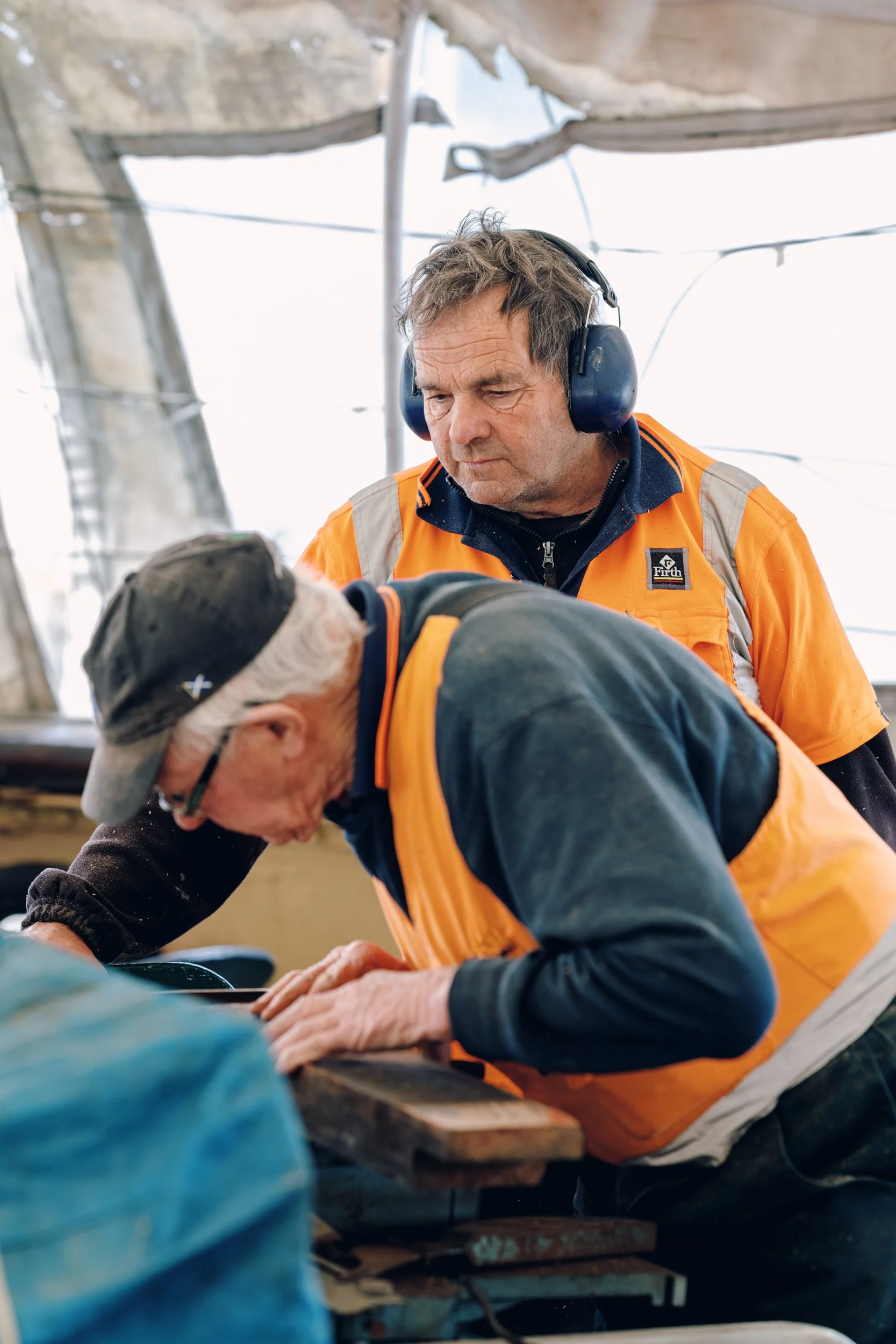 Two men working together indoors, with one wearing ear protection and the other taking a close look at a wooden surface.