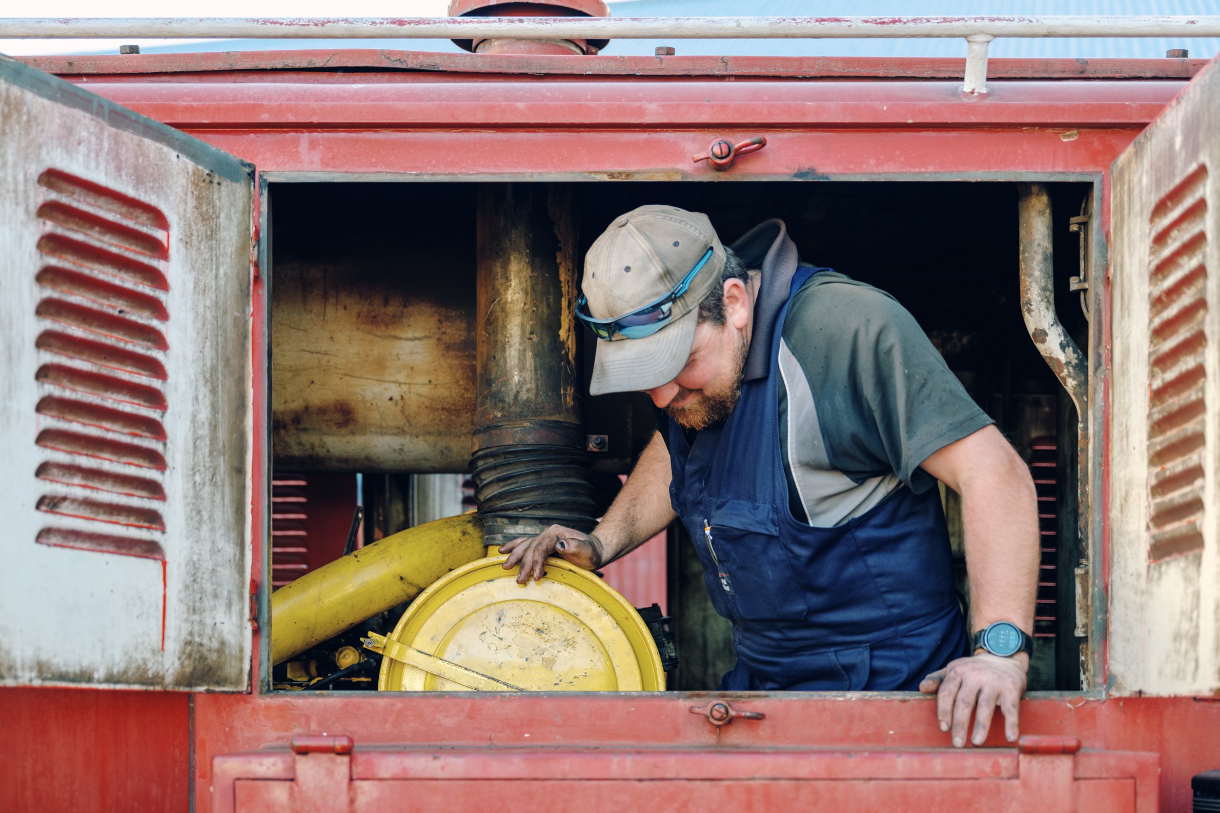 A man working on machinery inside a red machine or vehicle, wearing a gray cap, sunglasses on his cap, a gray and black shirt, a blue vest, and a wristwatch. He is leaning into the machinery and appears focused on his work.