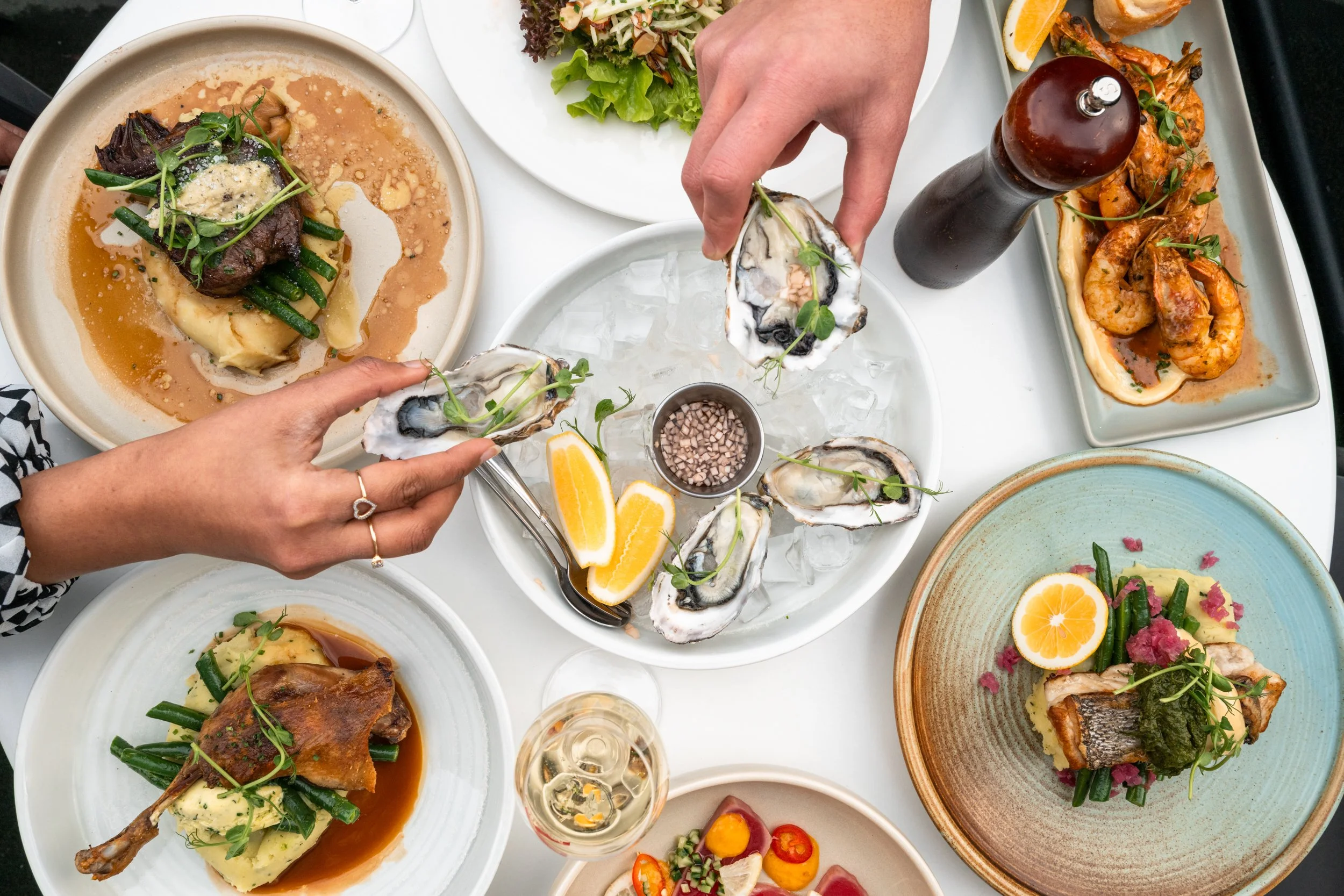 A variety of seafood dishes on a white table, including oysters on ice with lemon wedges, shrimp in a sauce, cooked fish with green beans and mashed potatoes, a lamb chop with gravy and vegetables, and a dessert with a lemon slice and berries, with a