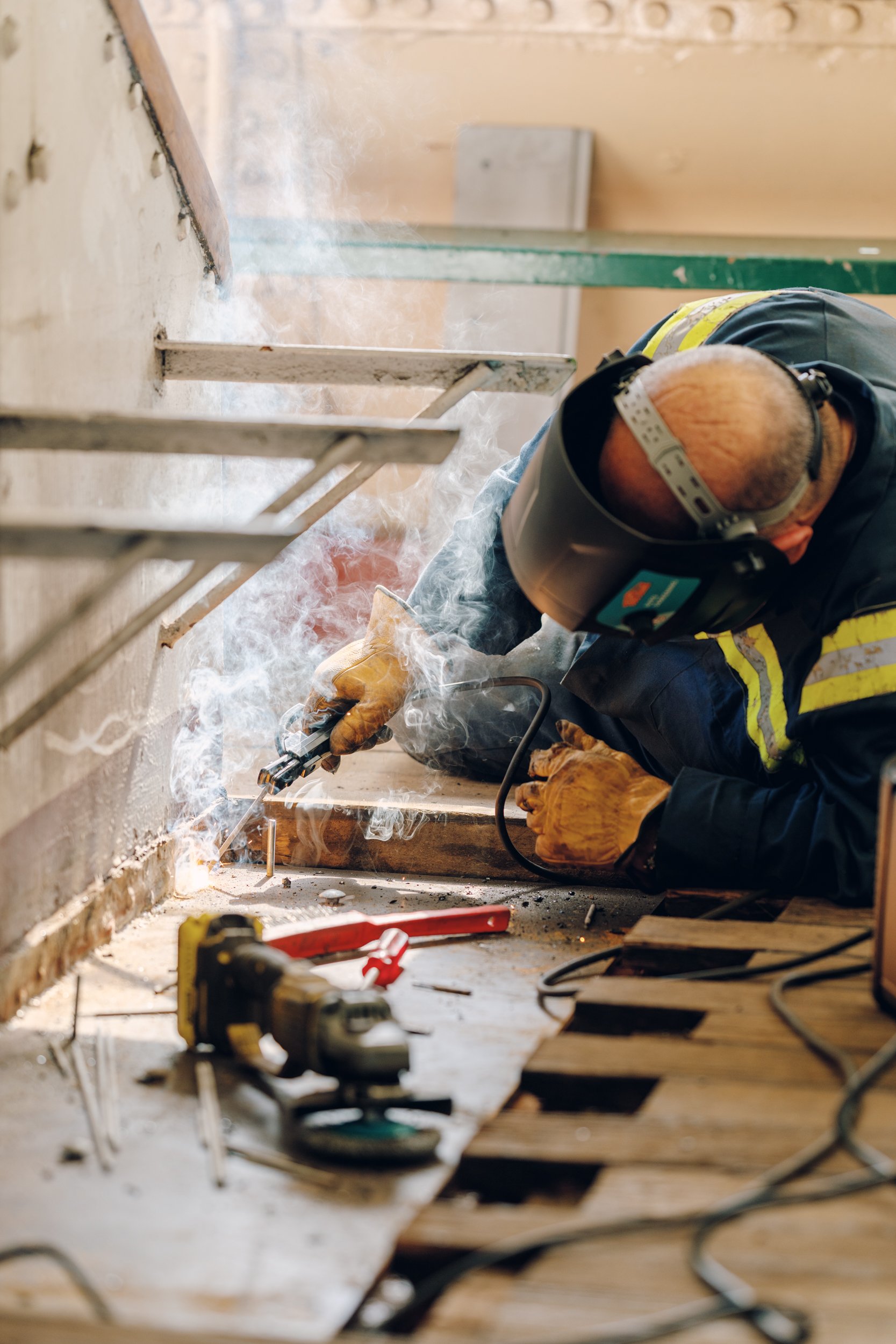 A construction worker welding metal pieces together on a wooden floor, wearing protective gear, gloves, and a welding helmet, with tools and equipment nearby.