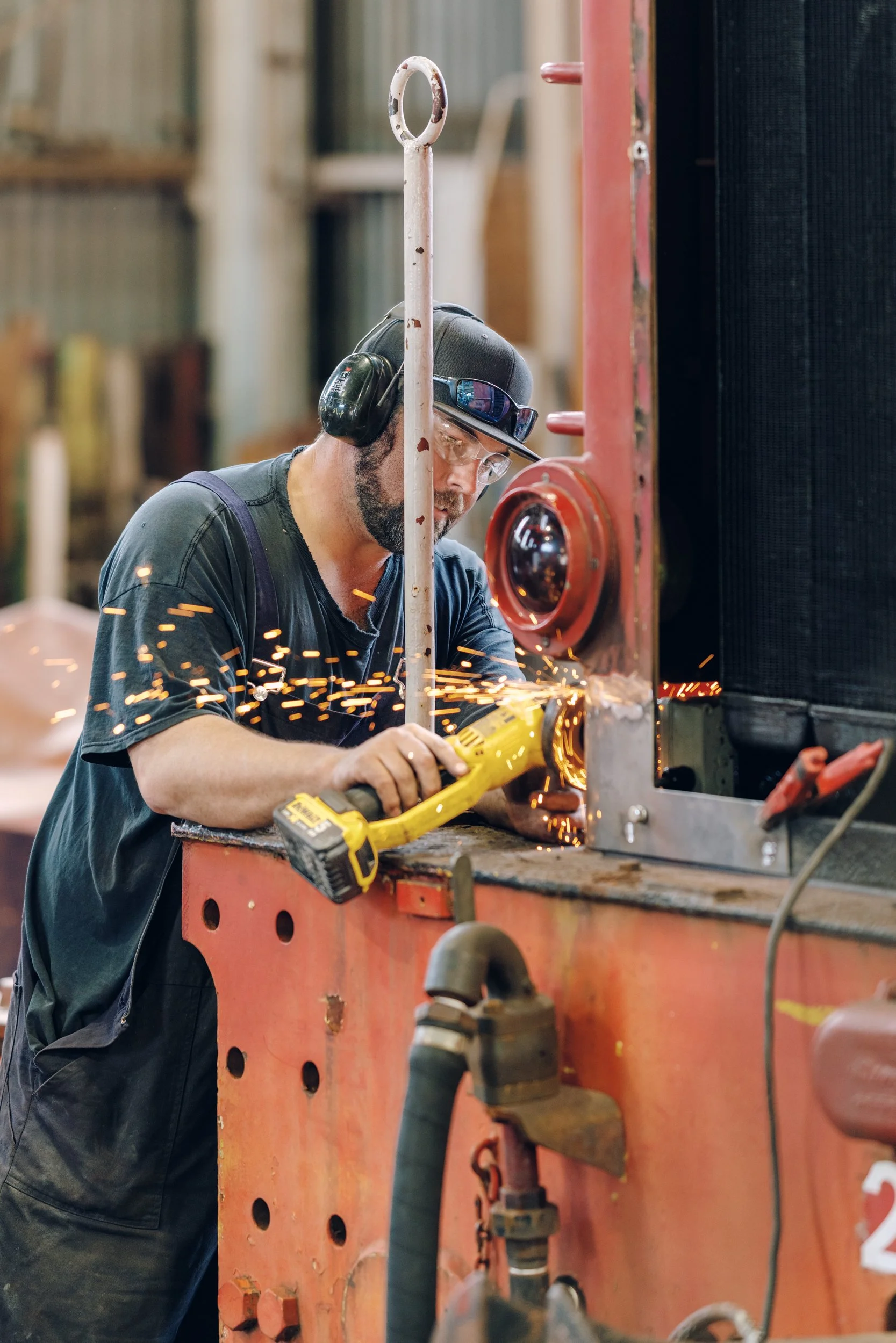 A man wearing safety glasses, headphones, a cap, and dark work clothes welding metal while sparks fly around him on an industrial workshop setting.
