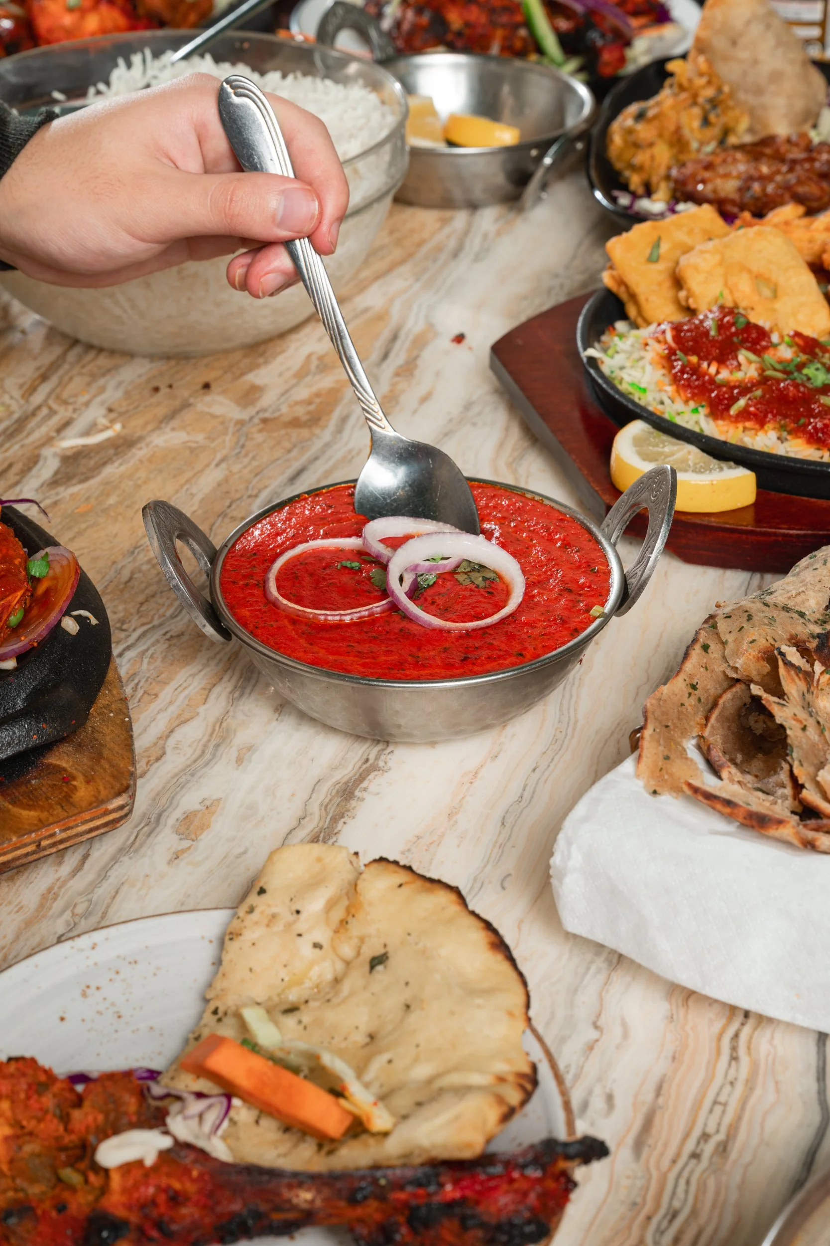 A small bowl of bright red tomato-based curry or sauce with onion slices on top, surrounded by various platters of Indian food including naan bread, rice, fried snacks, and grilled meat, on a marble tabletop.