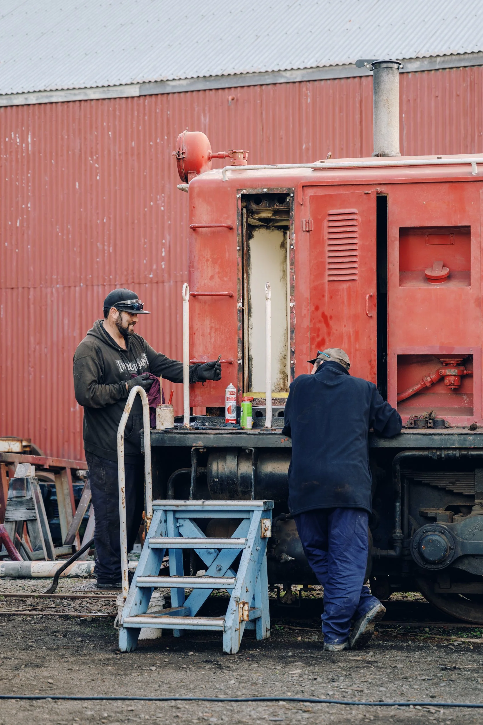 Two men working on an old red train engine outdoors, one standing and smiling, the other leaning over, with tools and supplies nearby.