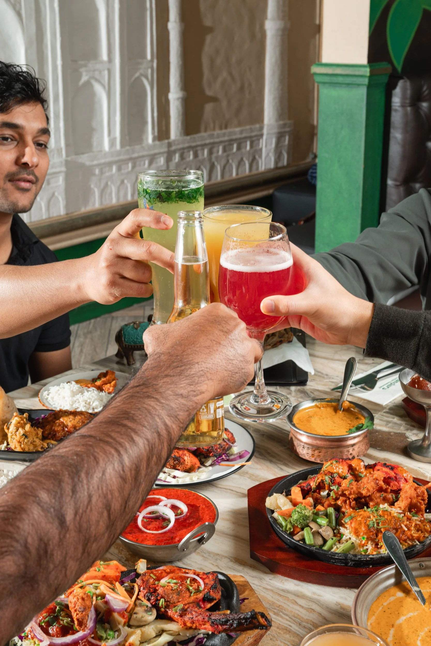 People toast with colorful drinks over a table filled with Indian dishes and condiments at a restaurant.