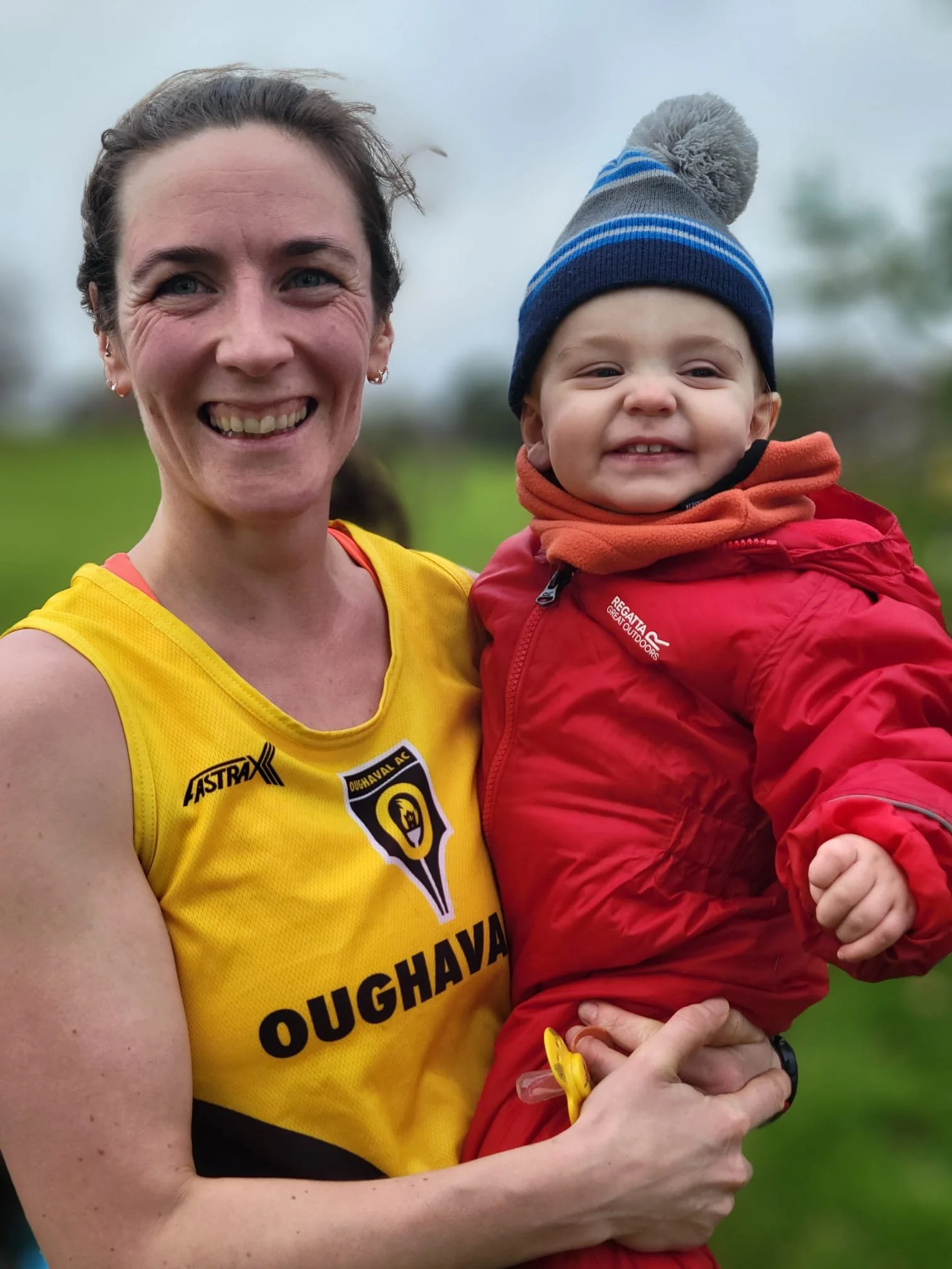 A smiling woman with short dark hair holding a young child dressed in a red jacket and a blue striped beanie hat outdoors.