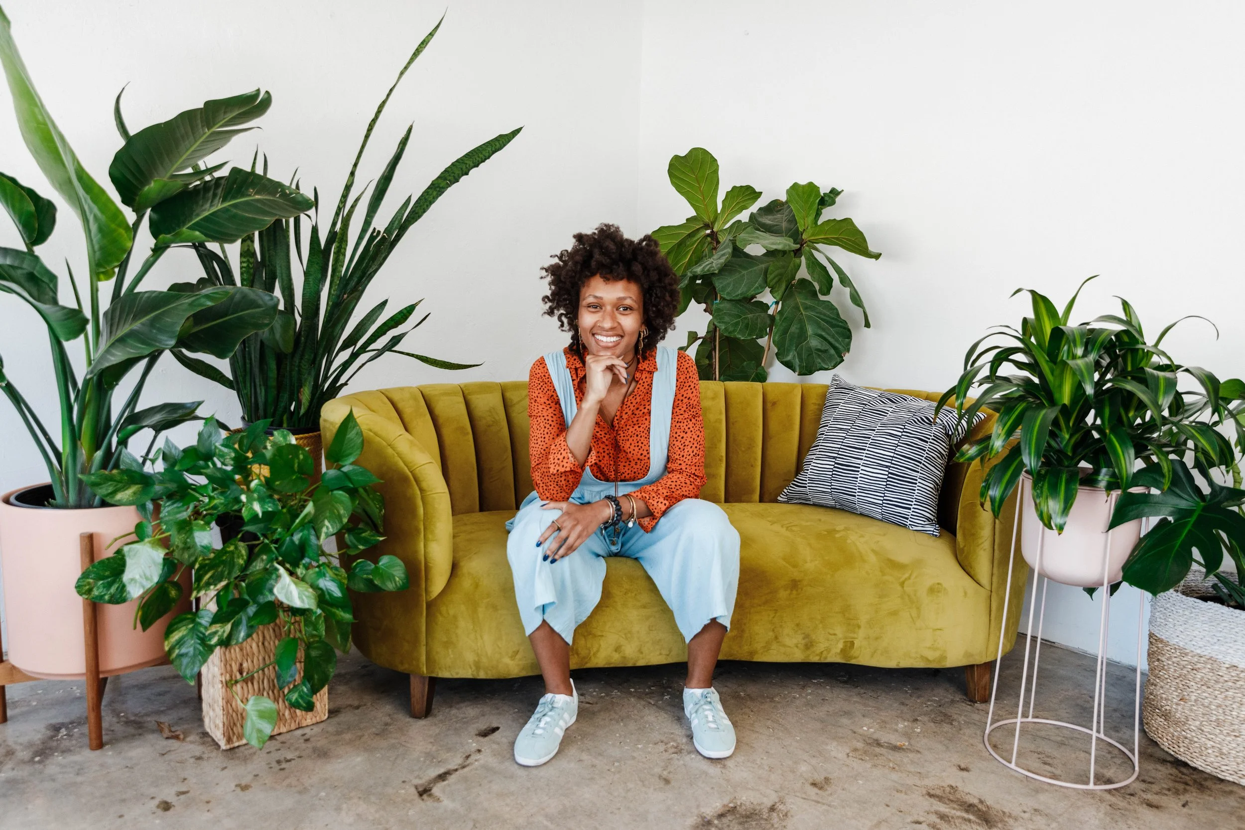 You Belong Here Coworking Space Manager Elisa, a brown-skinned woman with curly hair, sits on a couch surrounded by plants.