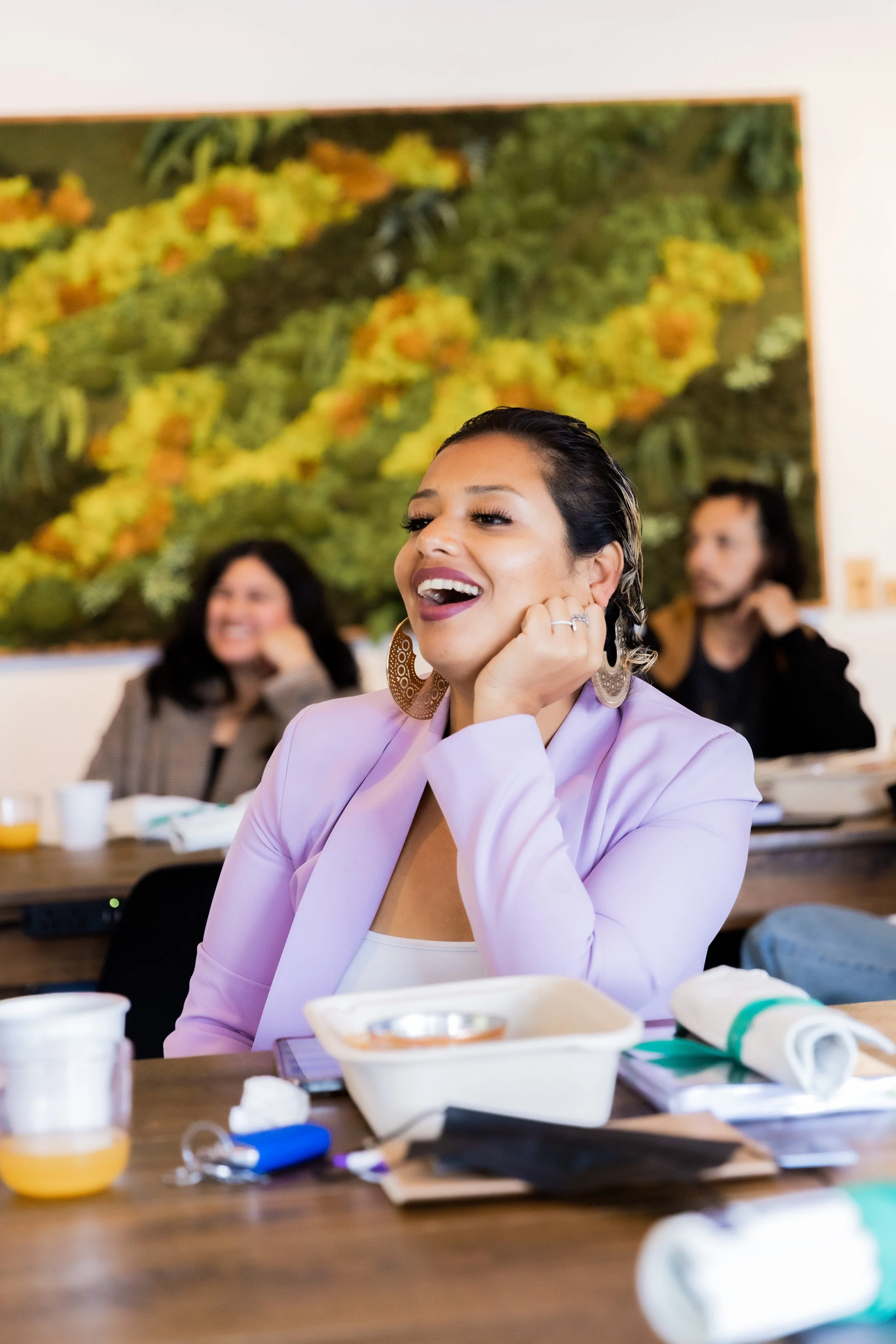 Brenda, a You Belong Here member with light brown skin and dark hair, laughing while seated with other community members behind her.