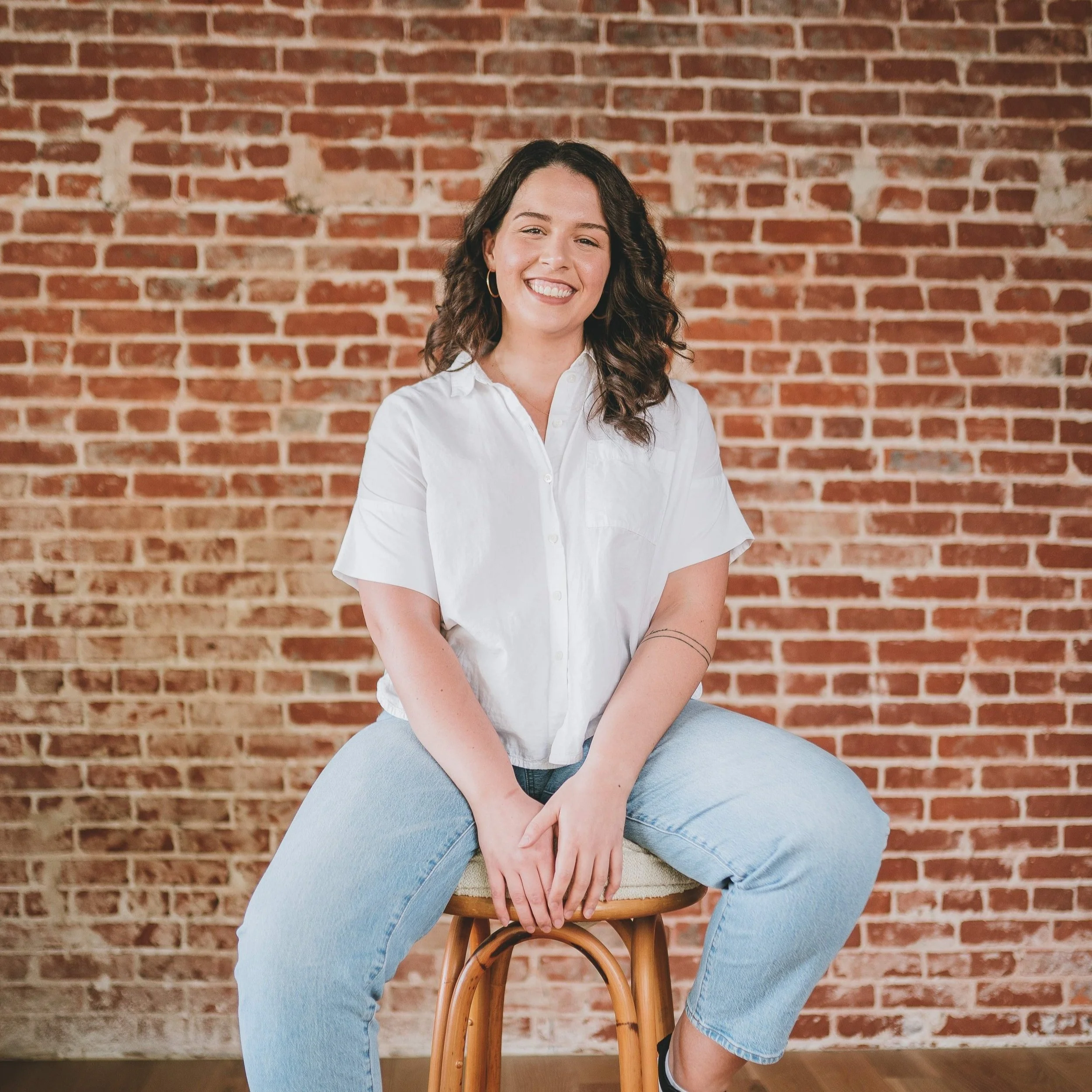 You Belong Here Blog Contributor Hannah, a white person with brown hair, sits on a stool in front of a brick wall.