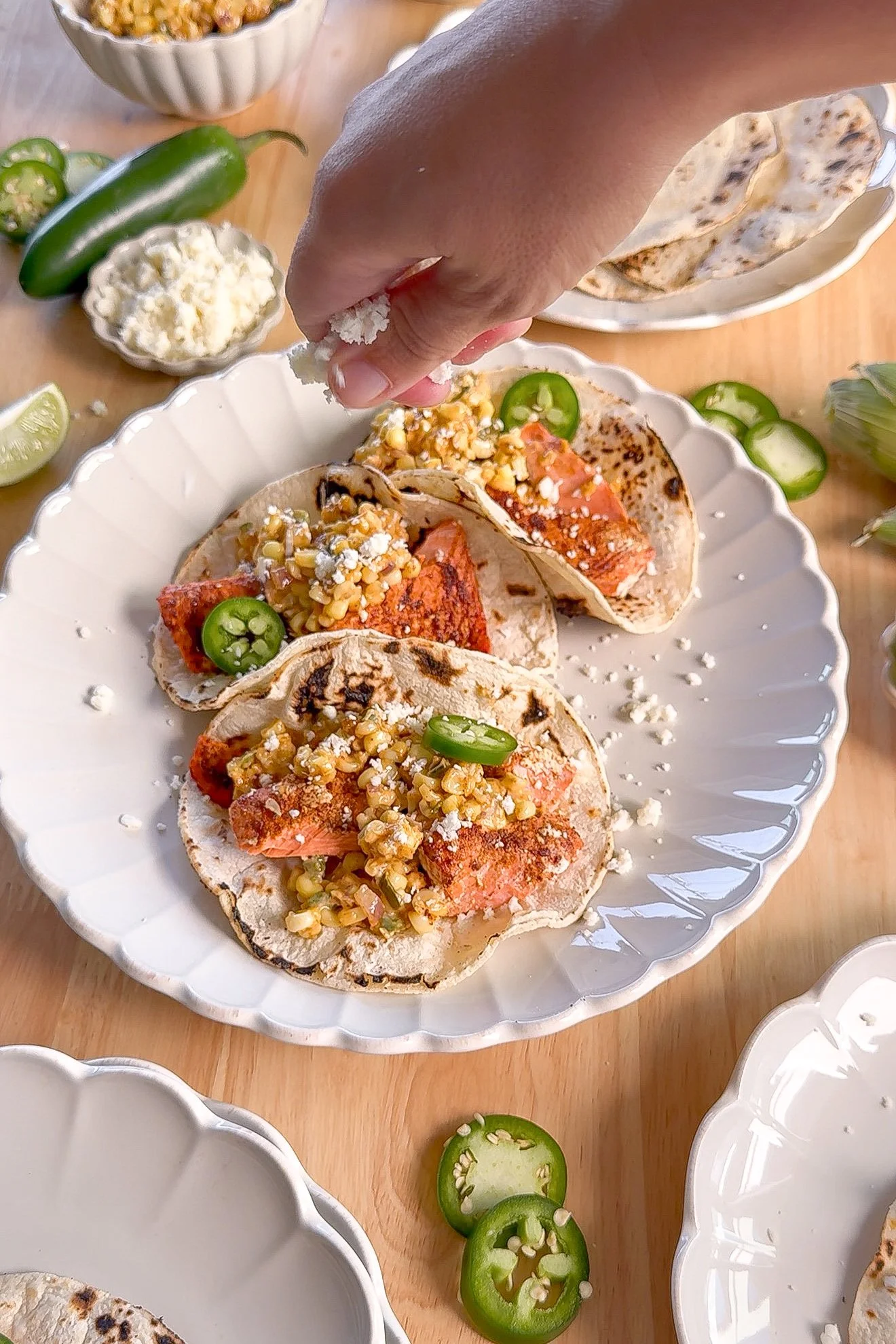 Crumbled cotija cheese being sprinkled over salmon tacos topped with creamy elote corn salsa.