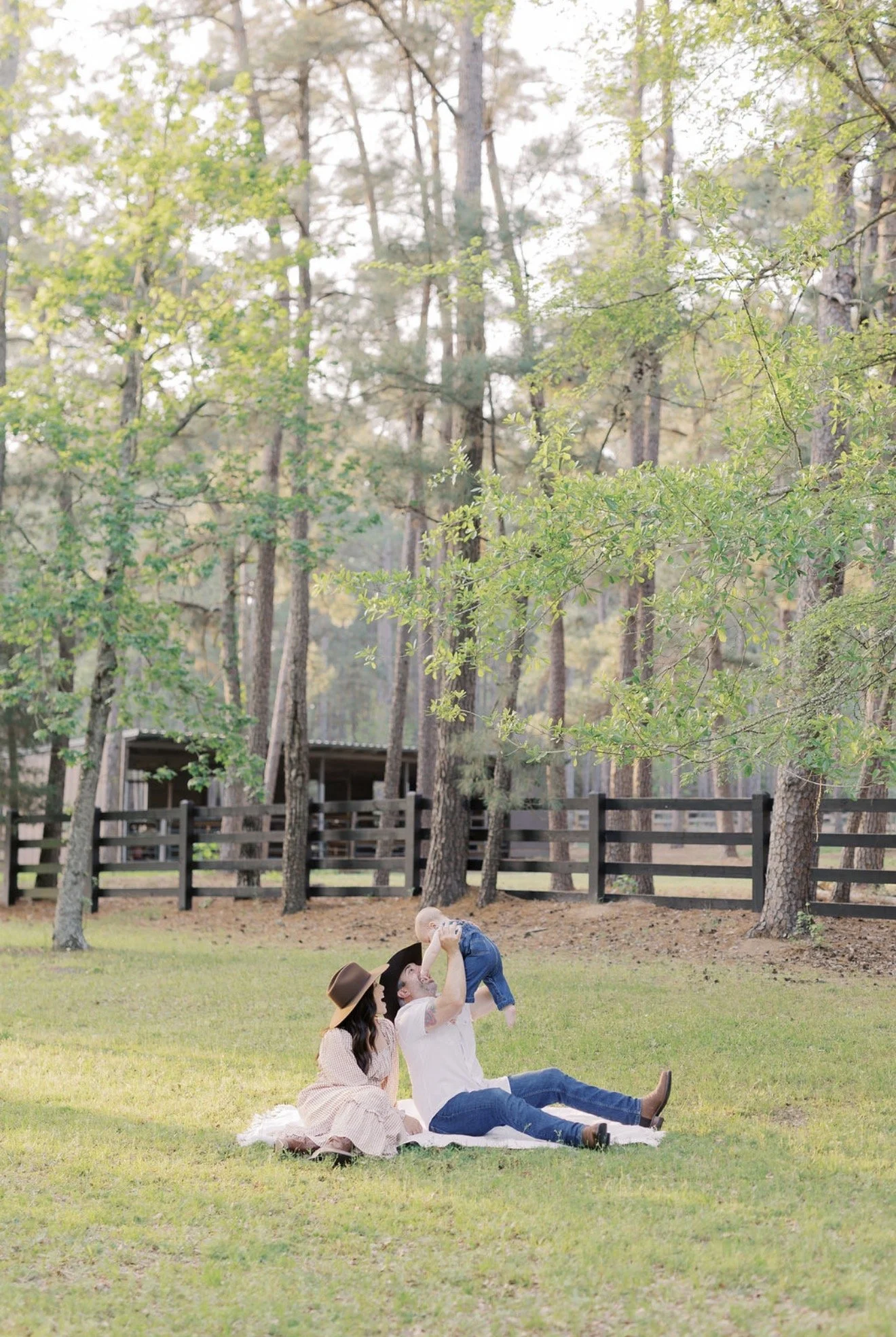 Mom and dad sitting on a blanket as dad holds their baby in the air with their Texas acreage behind them