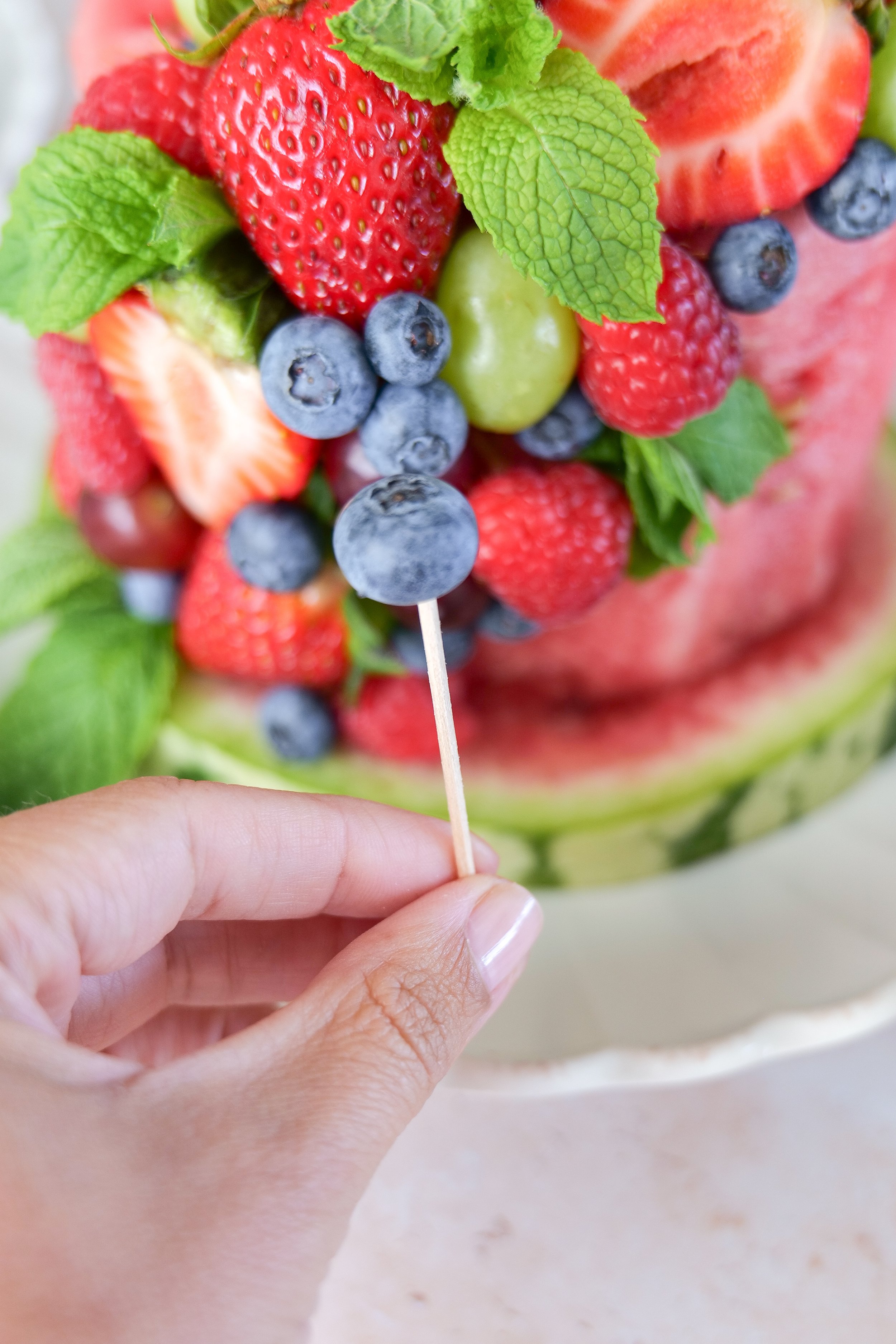 Fresh berries and grapes being attached to a watermelon cake with toothpicks for a colorful summer dessert.
