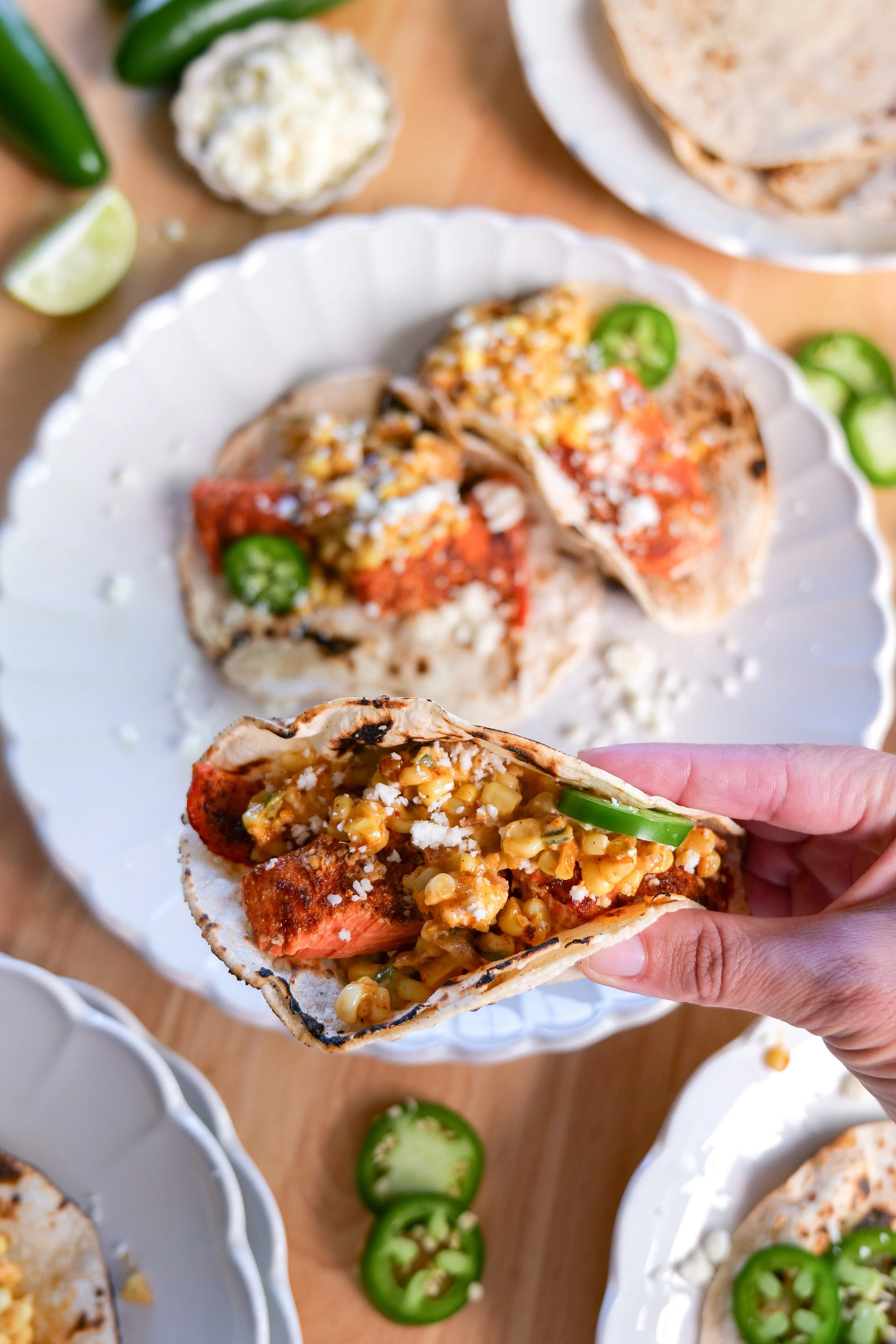 Close-up of a hand holding a homemade salmon taco filled with seasoned salmon, creamy elote corn salsa, and crumbled cotija cheese.